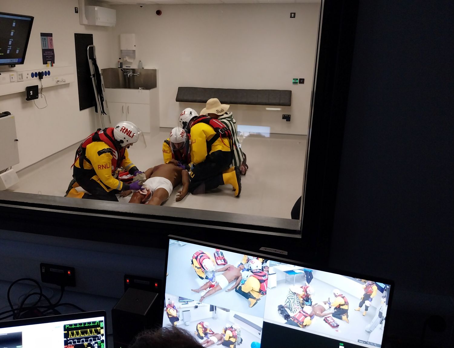 View of three crew in a clinical room carrying out a first aid exercise with a manikin and in the foreground the monitors of the control room are visible