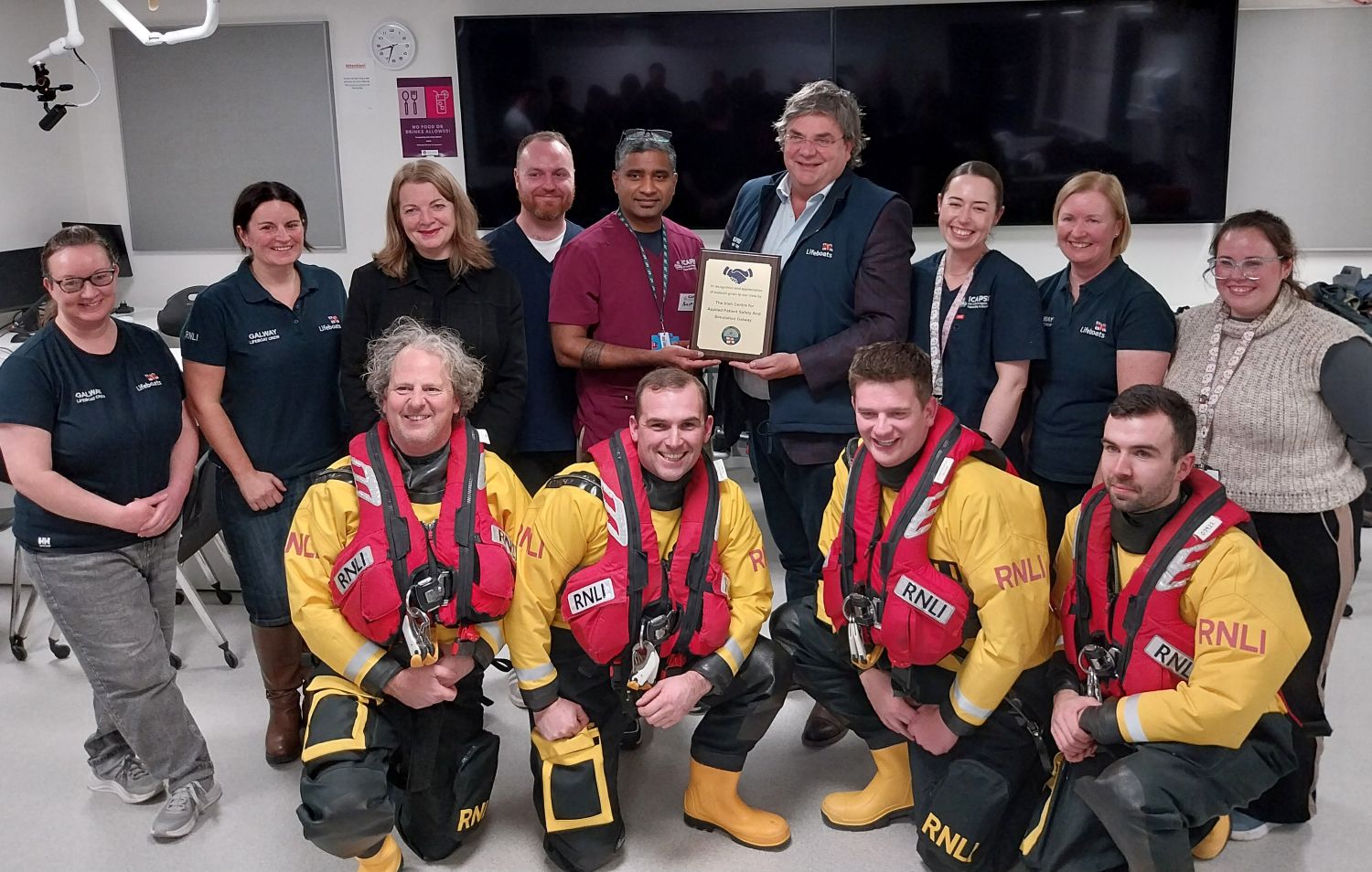 Four crew in drysuits kneeling in front of a group of nine standing. A plaque is being presented by RNLI to ICAPSS clinical staff