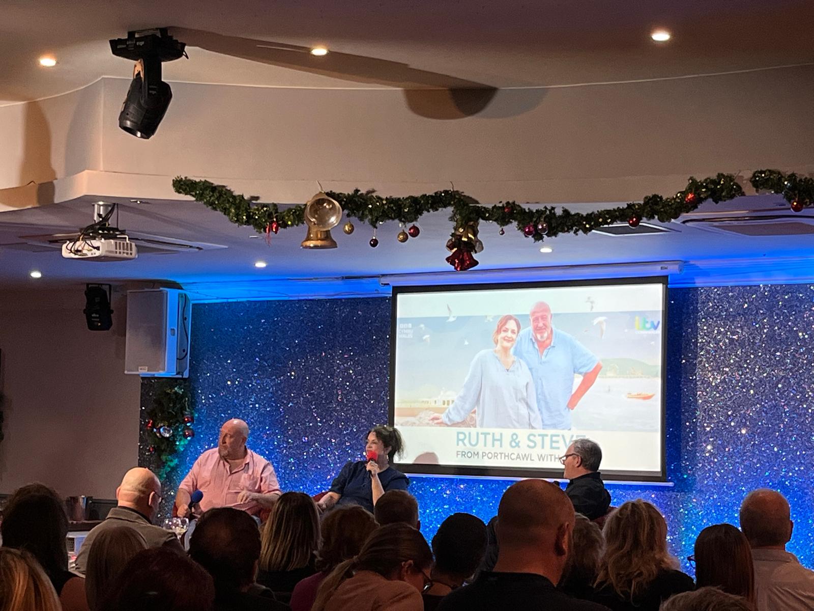 It's a room full of people with Ruth and Steve at the front on a stage and a big screen behind them with the opening of the programme.
