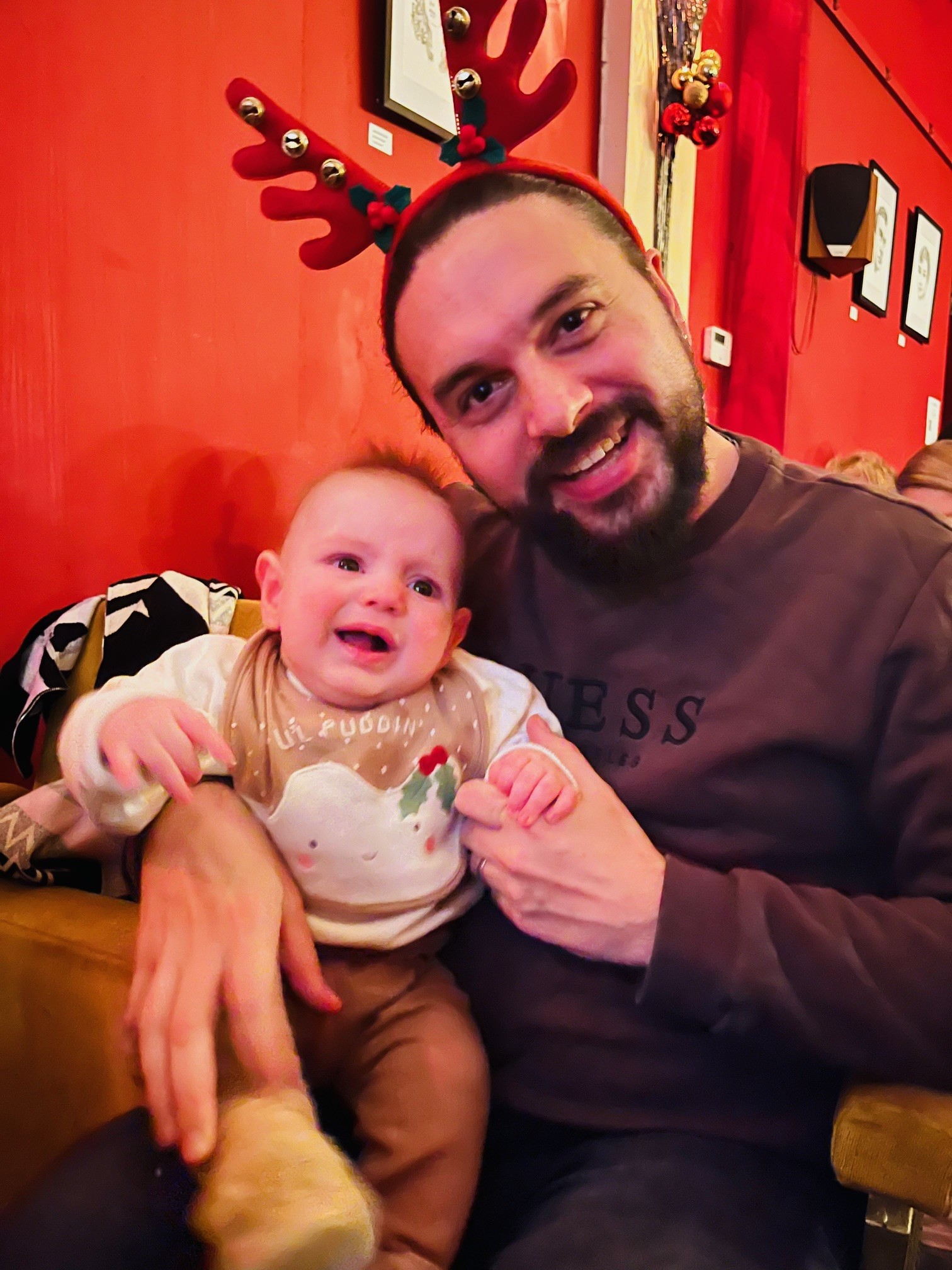 Yound father wearing a reindeer antlers head band holding his baby boy and smiling at the camera
