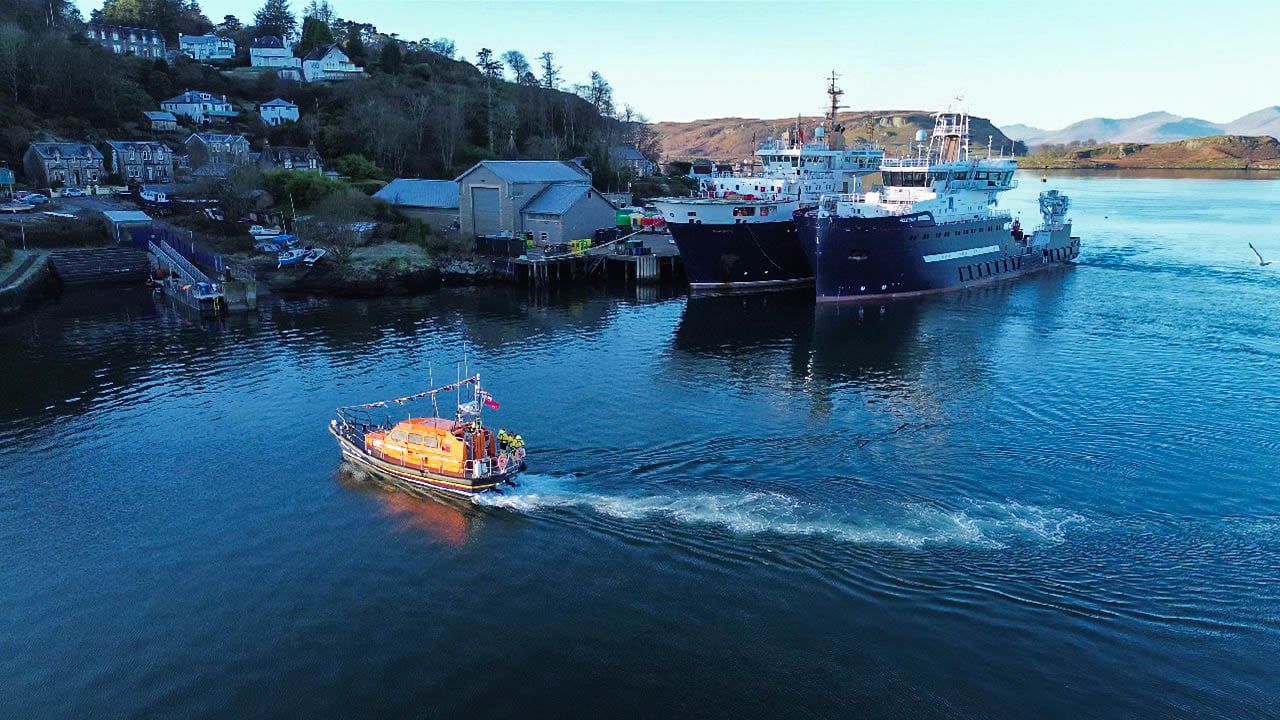 The lifeboat lit in the sunshine makes a sweeping arc in front of two moored lighthouse support ships.