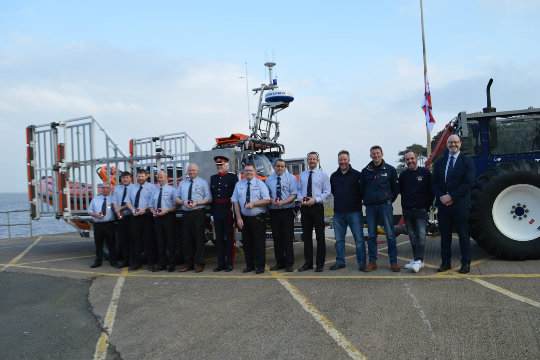 Crew members standing infront of orange lifeboat holding their medals, deputy lord leitantant and RNLI Area Operations Manager also stood with crew members
