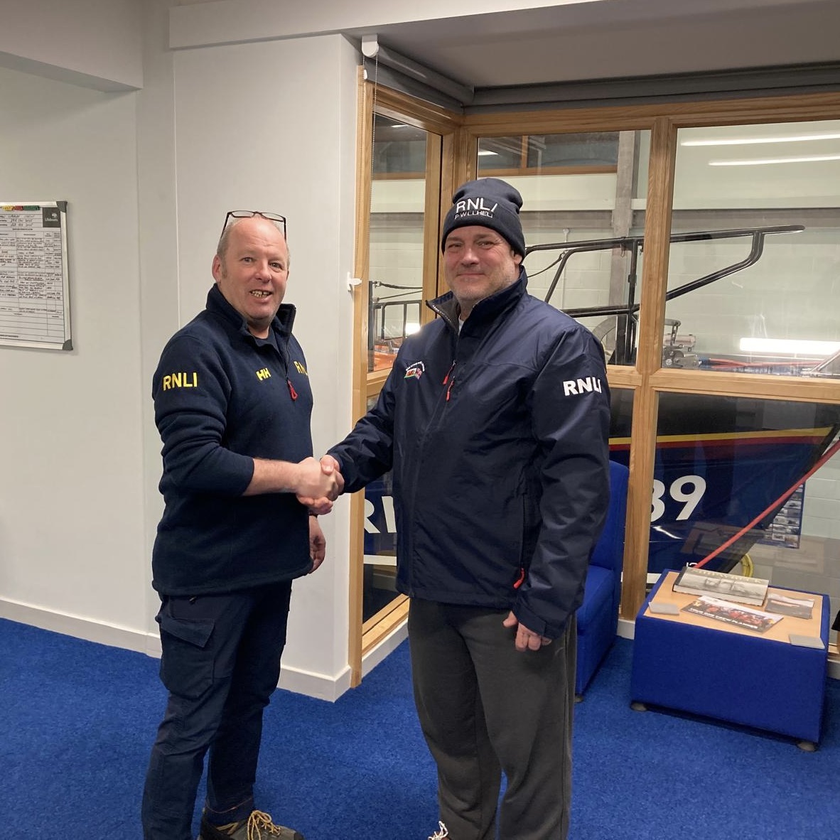 Simon Buntin on the left, shaking hands with Paul Kitteingham on the right. The all-weather lifeboat can be seen in the background through the windows.