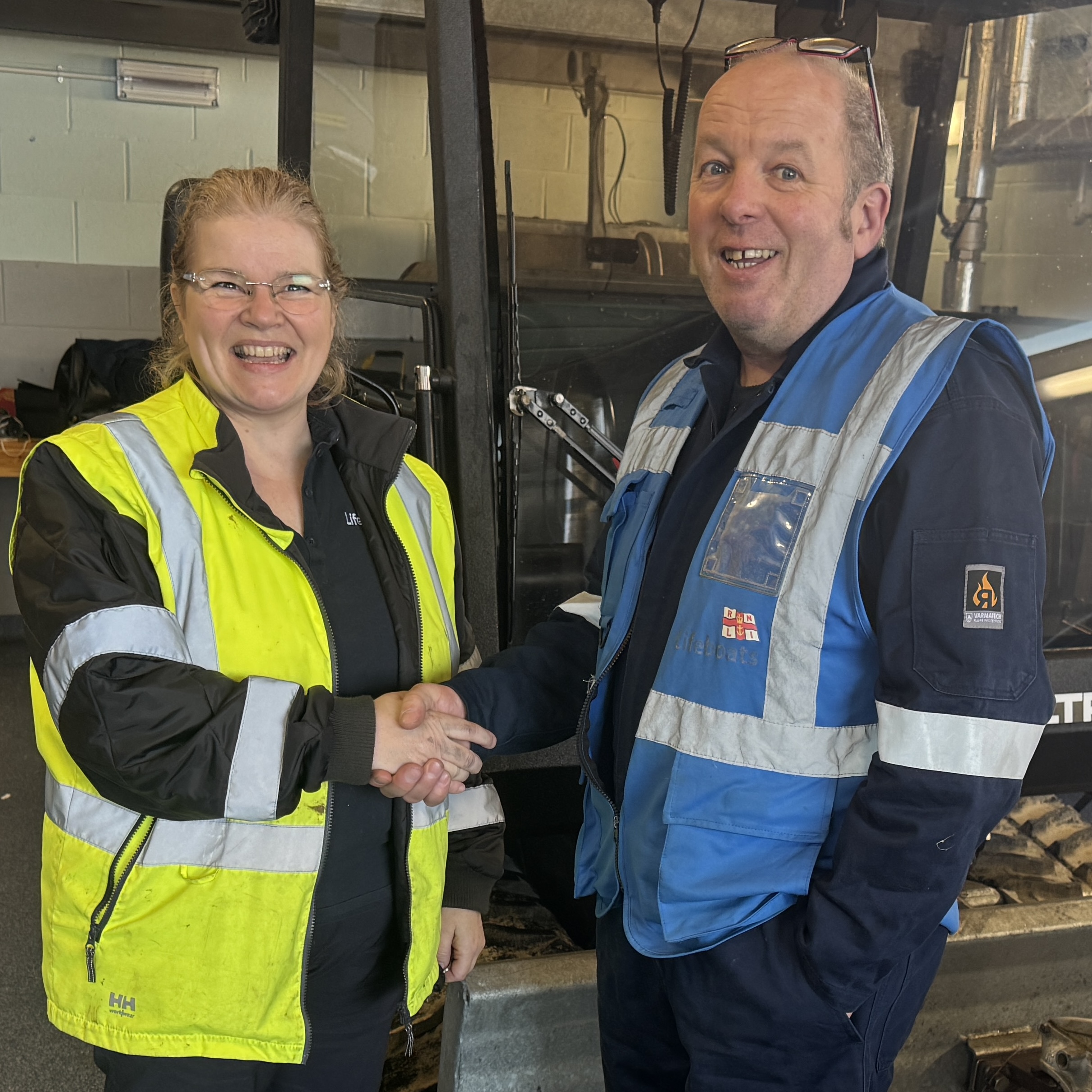 Caroline Jones on the left wearing RNLI high vis jacket, shaking hands with Simon Bunting, RNLI assessor. They are standing in front of a ToolTrack tracked launch vehicle.