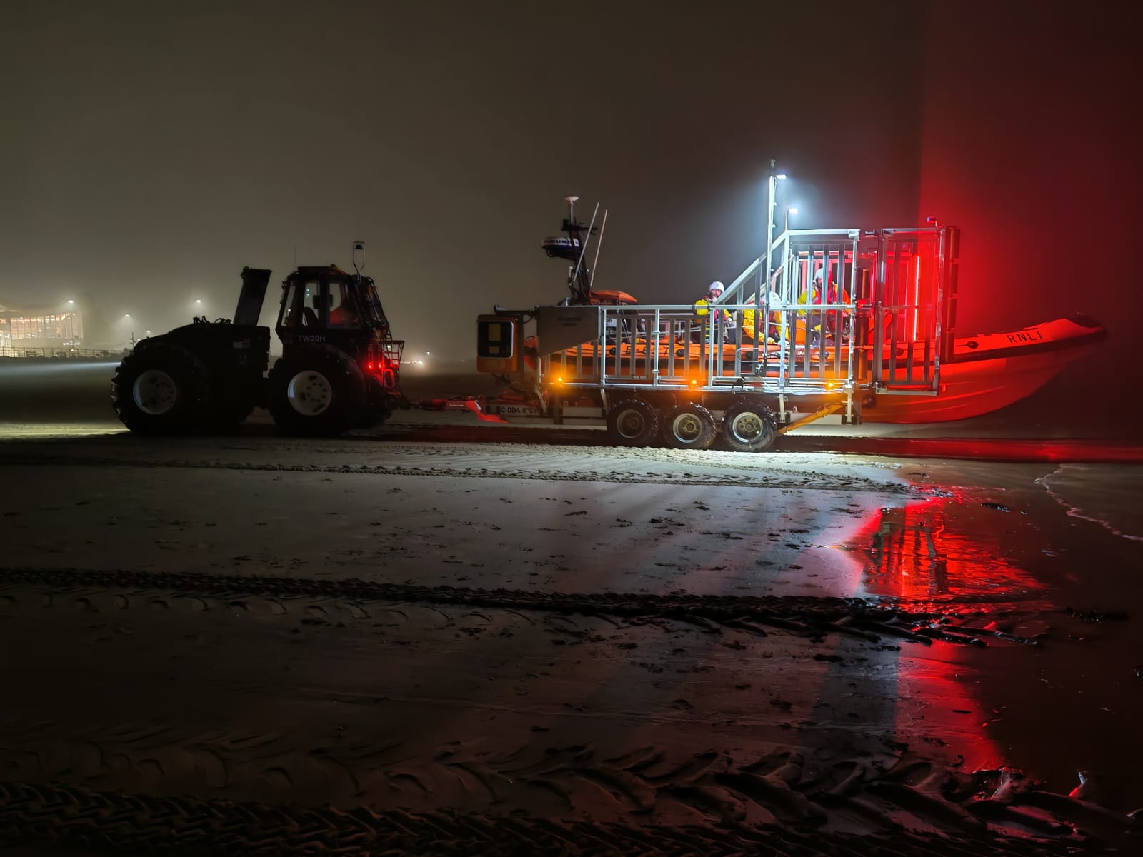 A dark night image, with New Brighton's tractor, carriage and lifeboat making their way to the launch site. Red and white lights shine on the slight fog.
