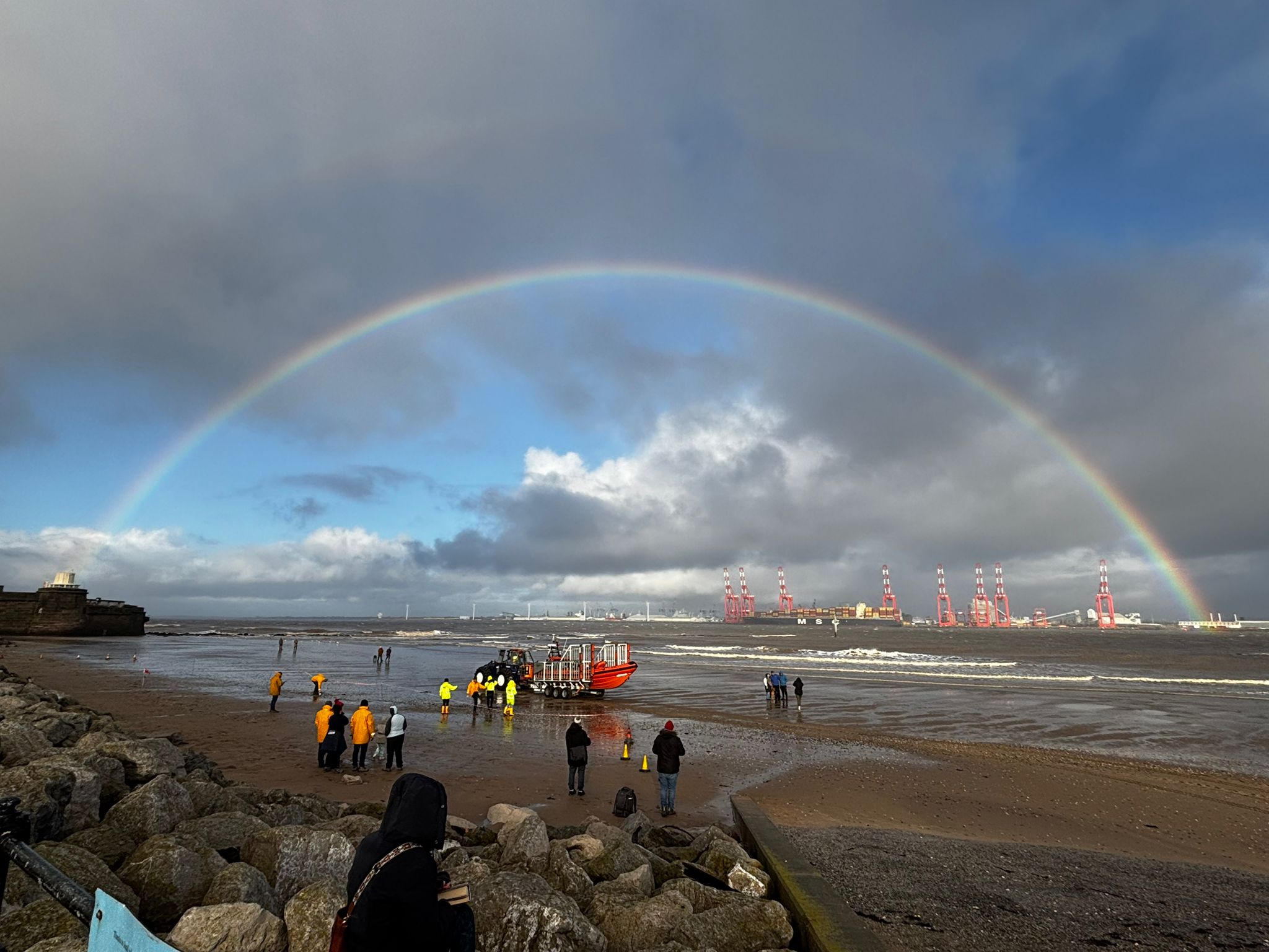 Landscape image of New Brighton beach. Volunteer crew in yellow can be seen on the sand, with the tractor, carriage and lifeboat just beyond them. A rainbow stretches across the sky, from the Fort on the left of the frame, to the Liverpool docklands on the right of frame. 