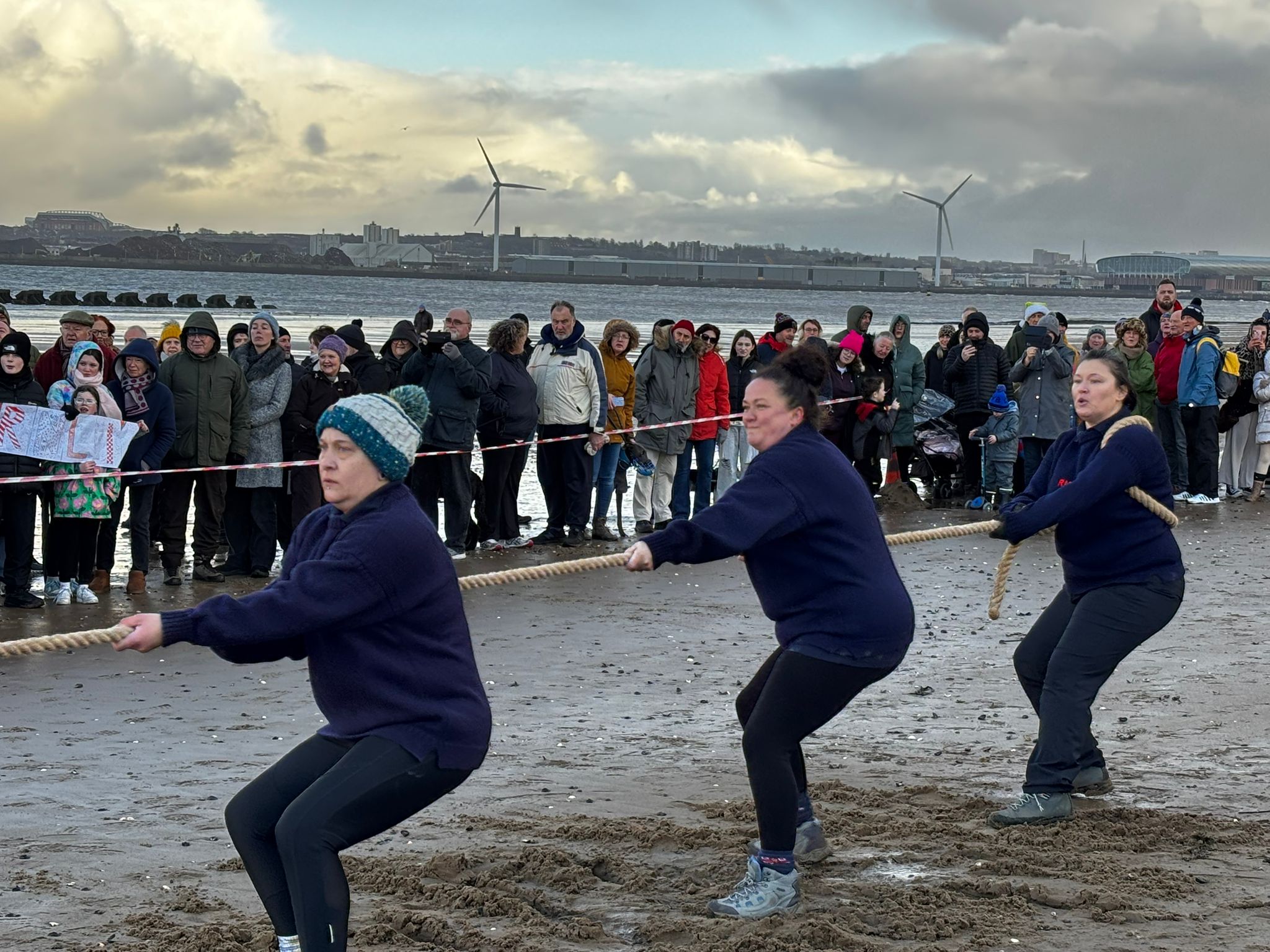 New Brighton beach. Three of New Brighton's women's team can be seen pulling hard on the tug of war rope. Behind the crowds of spectators can be seen watching on.