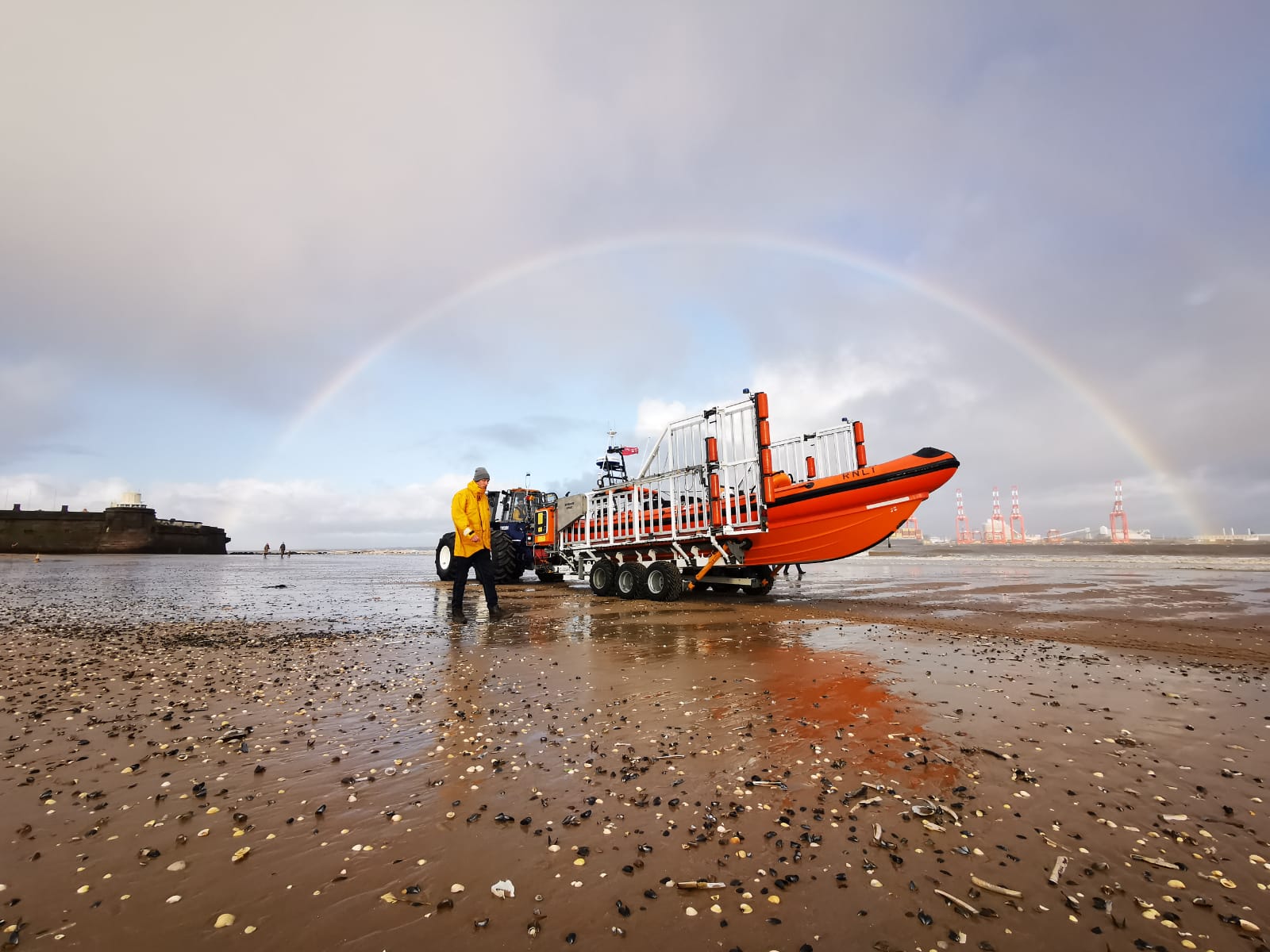 Low shot of New Brighton beach, with the Fort Perch Rock visible to the left of the frame. In the centre is the lifeboat on the carriage, with a single volunteer walking alongside. A rainbow stretches across the top of the carriage, tractor and lifeboat, framing it. 