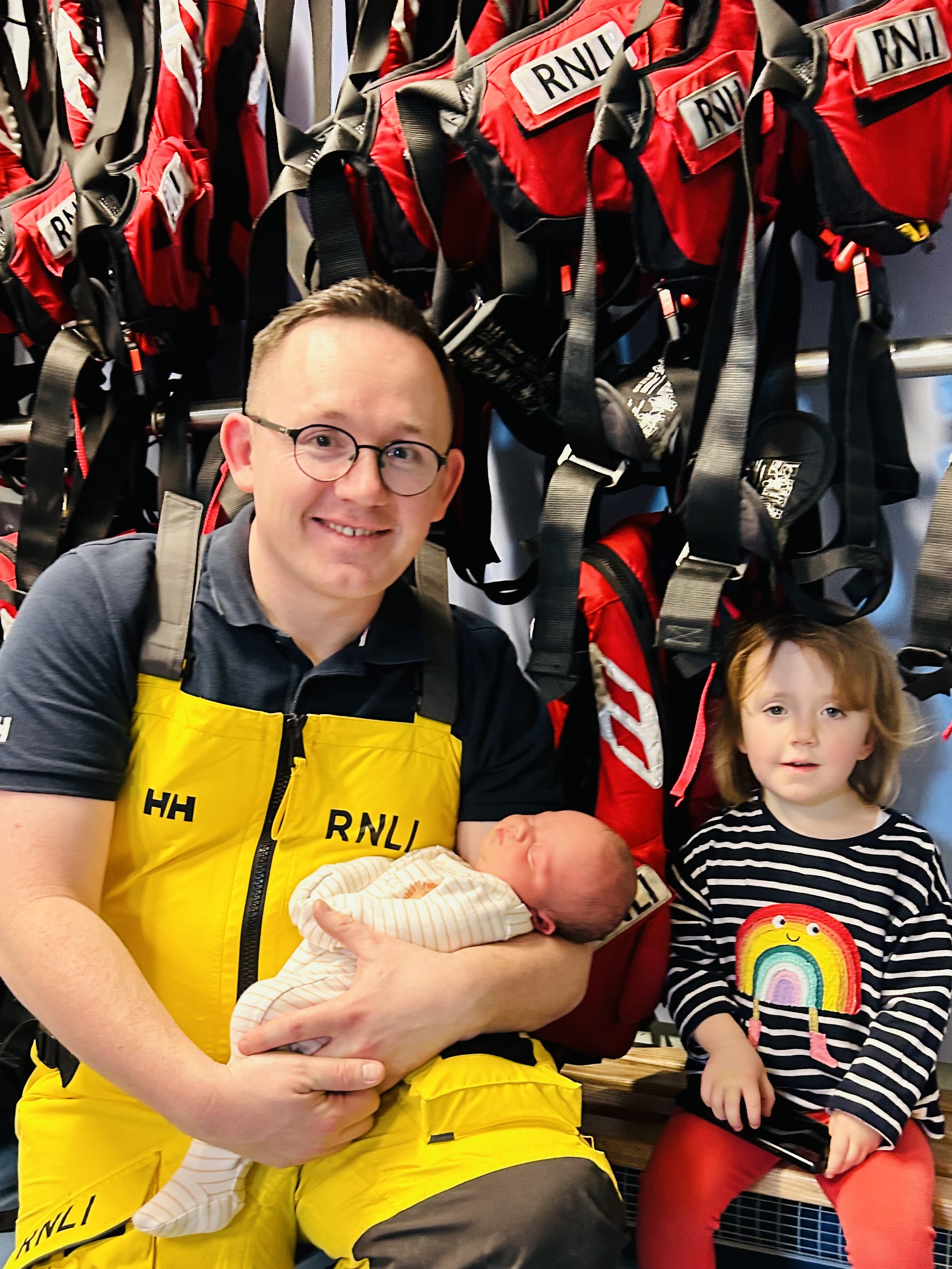 The photo shows a family of 3 - dad in RNLI yellows with his young daughter and baby son in front of some lifejackets