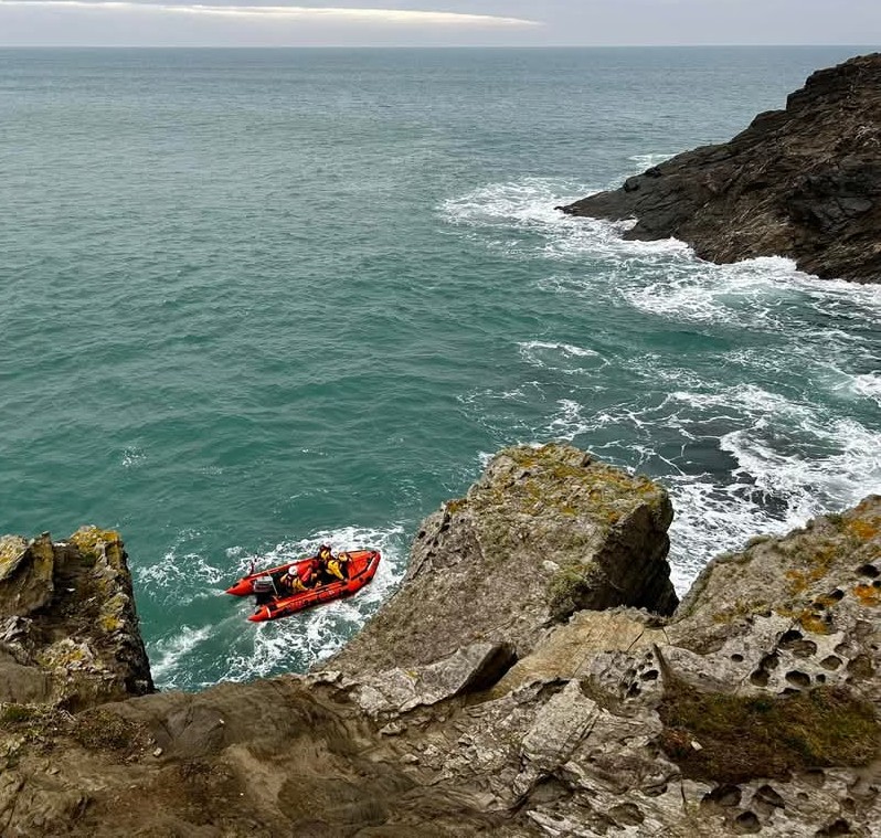View from top of cliff, with granite rocks in the foreground as Newquay inshore lifeboat searches white water around Porth Island