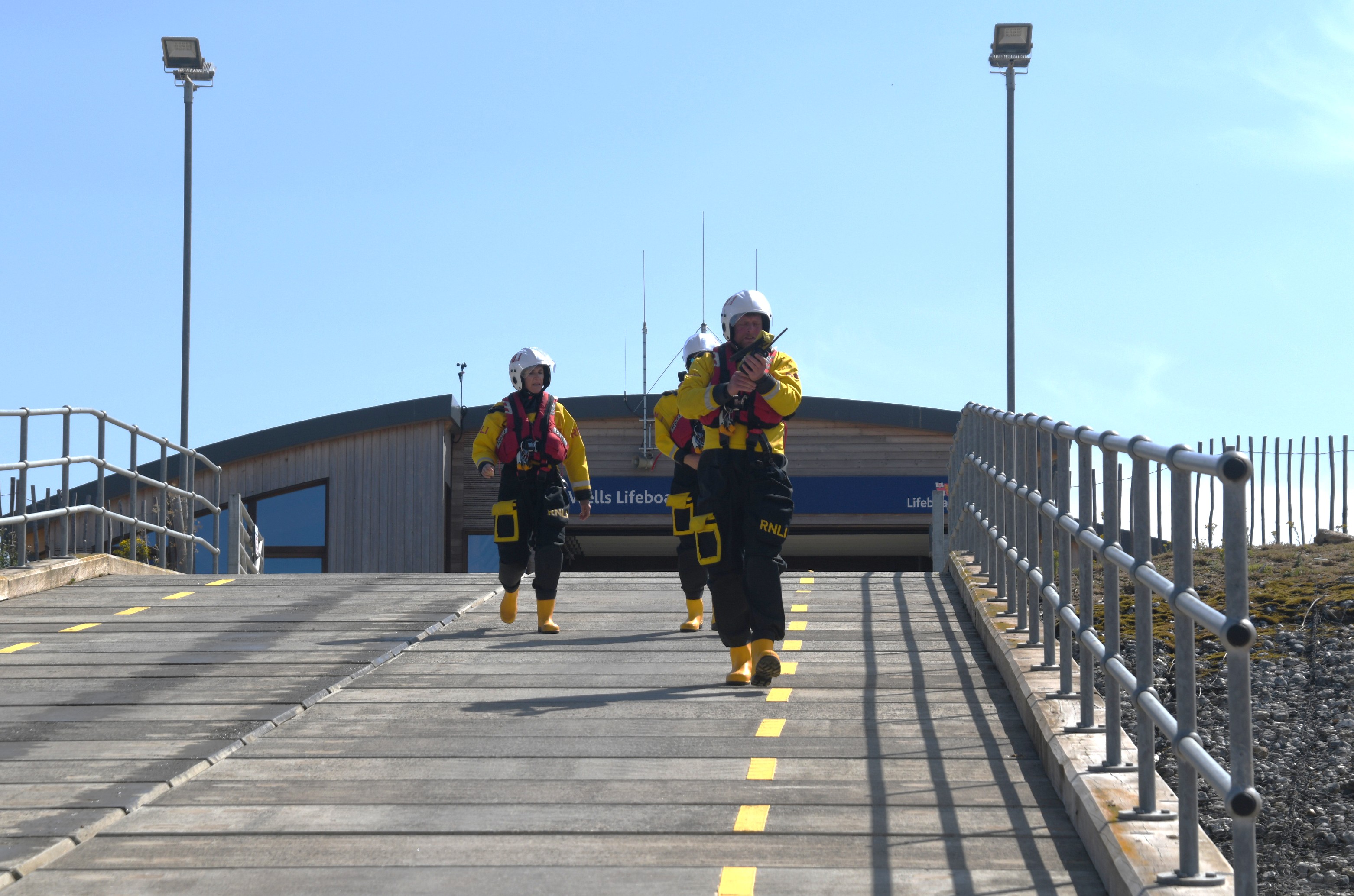 Three volunteer crew in full inshore lifeboat kit heading down slipway to launch. Lifeboat station in the background
