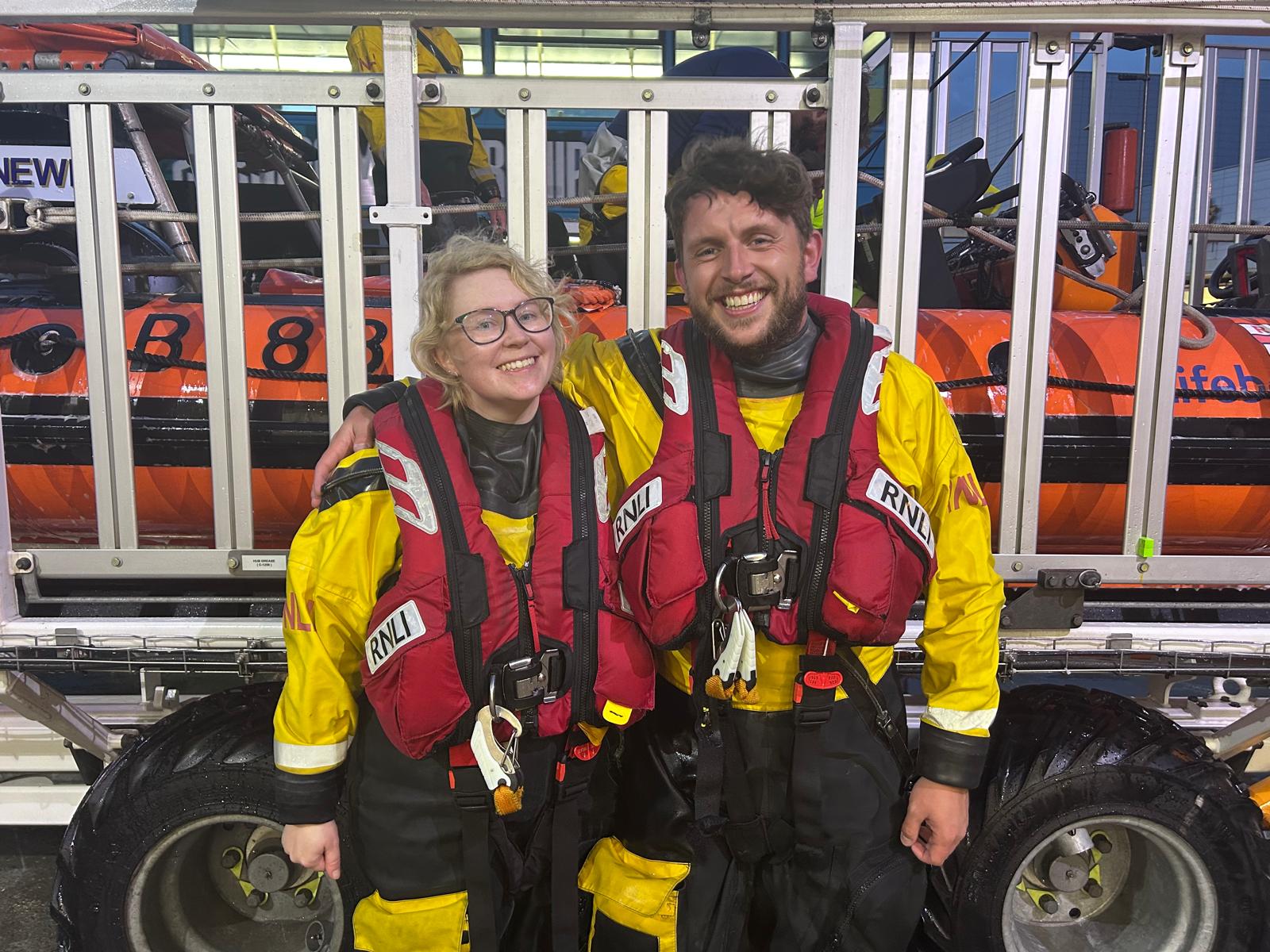 Steph and Connor pose in front of the lifeboat, on carriage. They are in drysuit and lifejackets and are all smiles.
