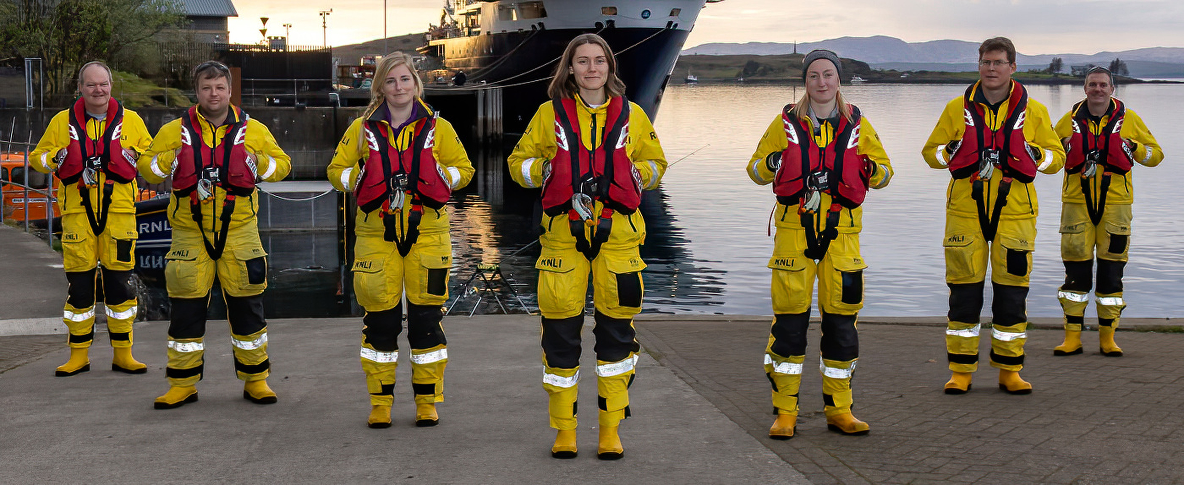 Seven men and women in their yellow all weather protective suits, yellow wellies and red lifejackets pose for a photograph on the quayside by Oban lifeboat station