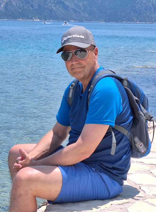A man wearing blue and a back back sits on a stone edge with sea and mountains in the background.