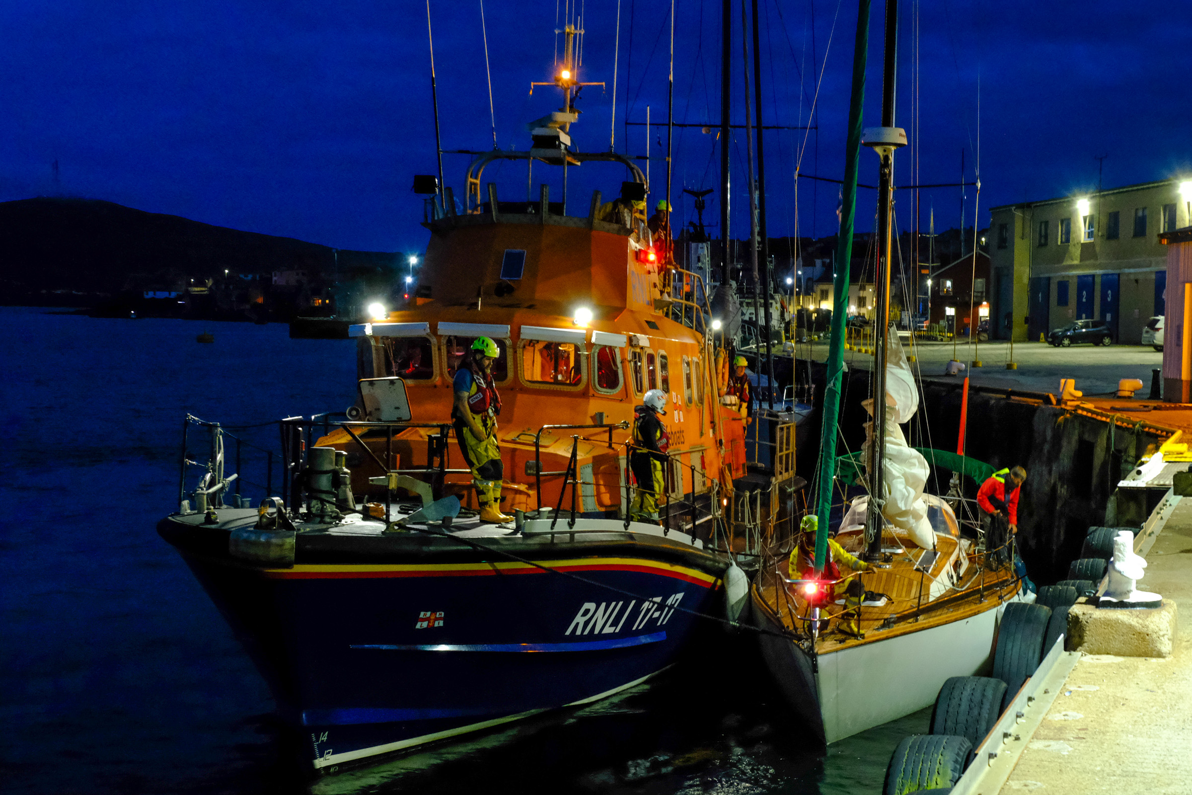 The lifeboat brings the yacht to a safe berth in Lerwick harbour - the image looks at the bow of both boats as crew pass lines and tie up the yacht