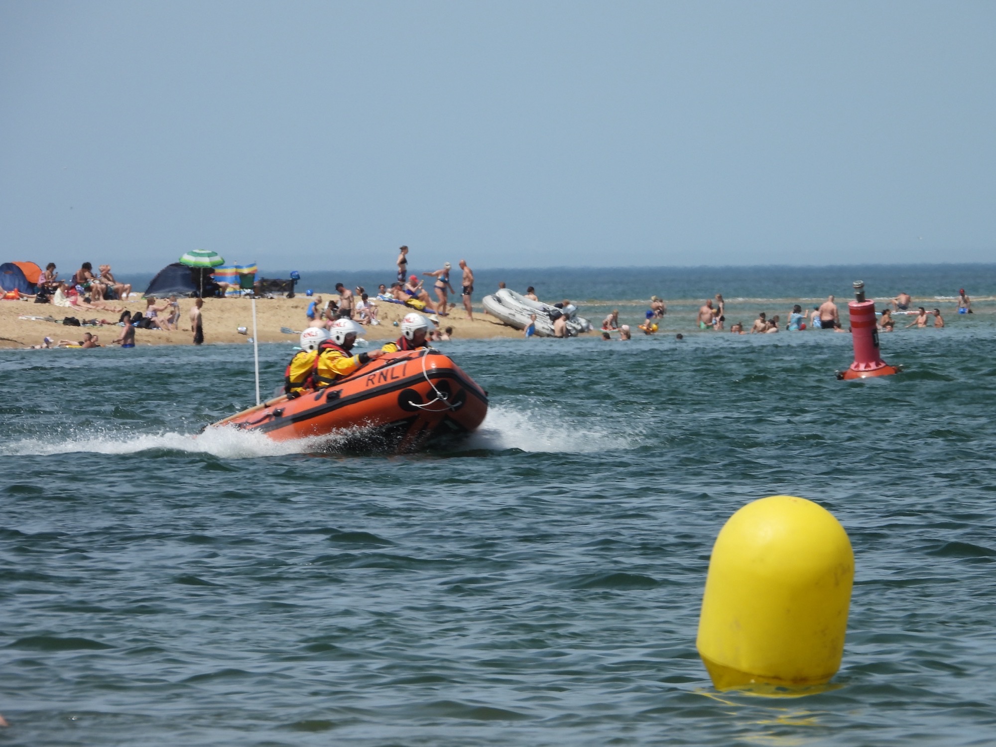 Wells RNLI Inshore Lifeboat Peter Wilcox on open water with people on the beach Three crew in inshore lifeboat kit and yellow and read bouys in water