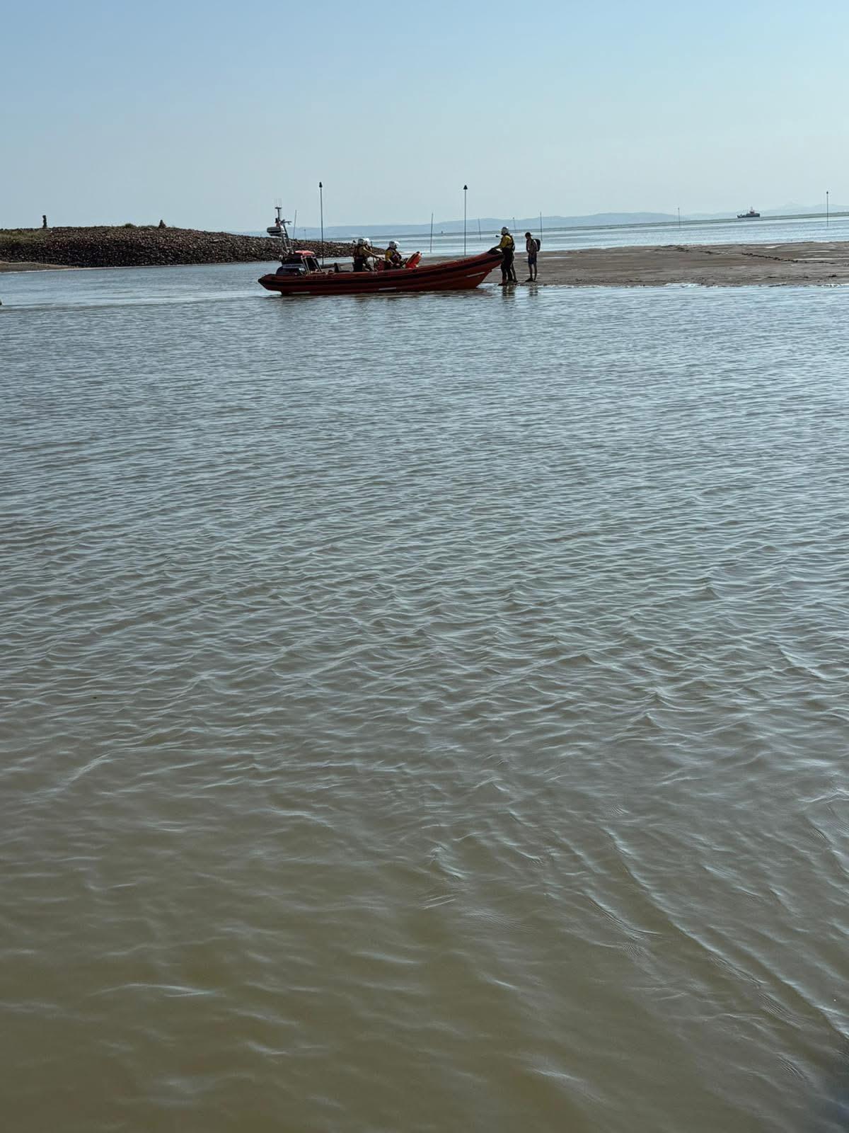 Portrait shot, primarily of water on the River Alt. At the top of the frame New Brighton RNLI's lifeboat can be seen, with two casualties about to climb aboard from the muddy bank.
