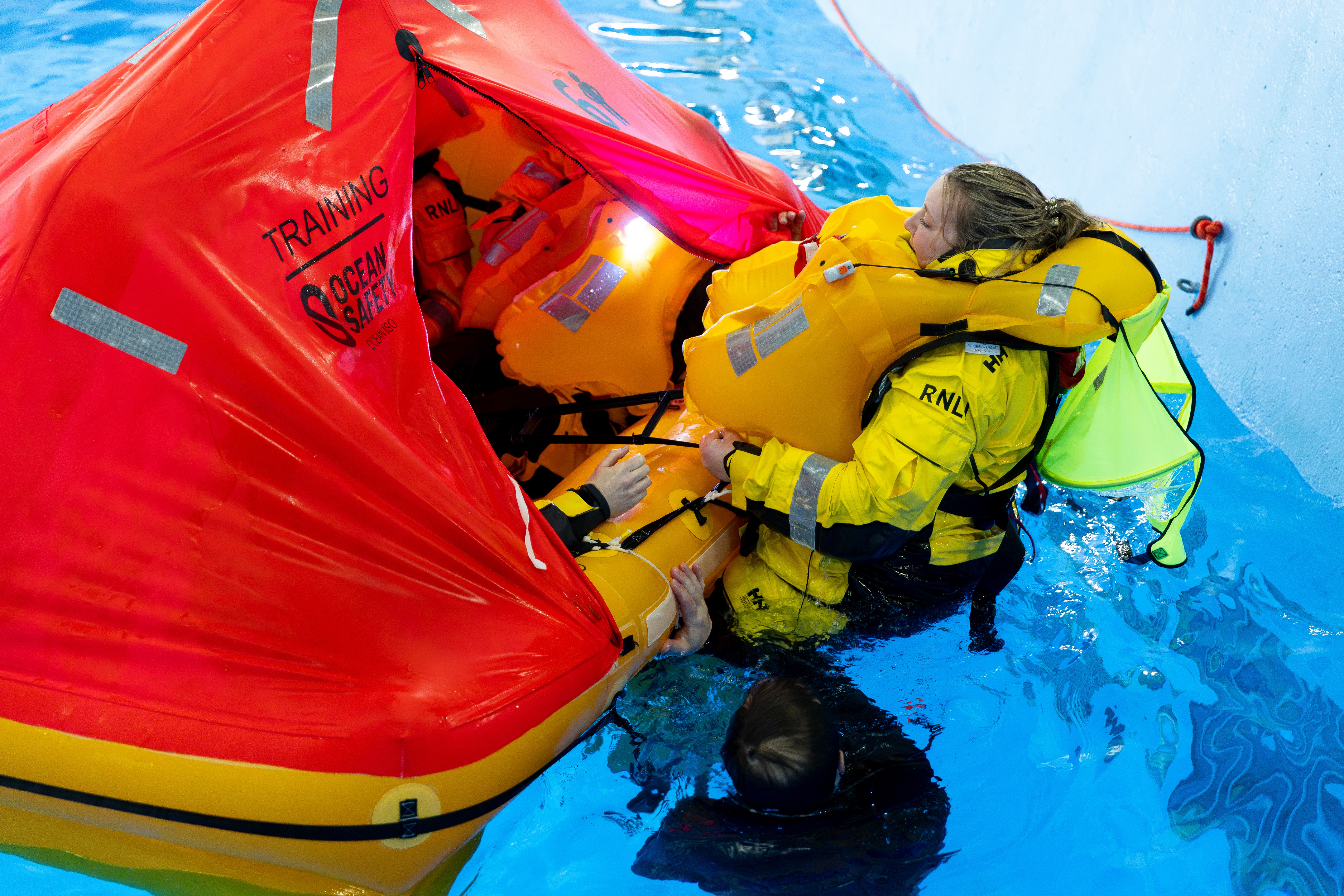 A woman in yellow kit and wearing a lifejacket climbs onto an orange life raft in the water.