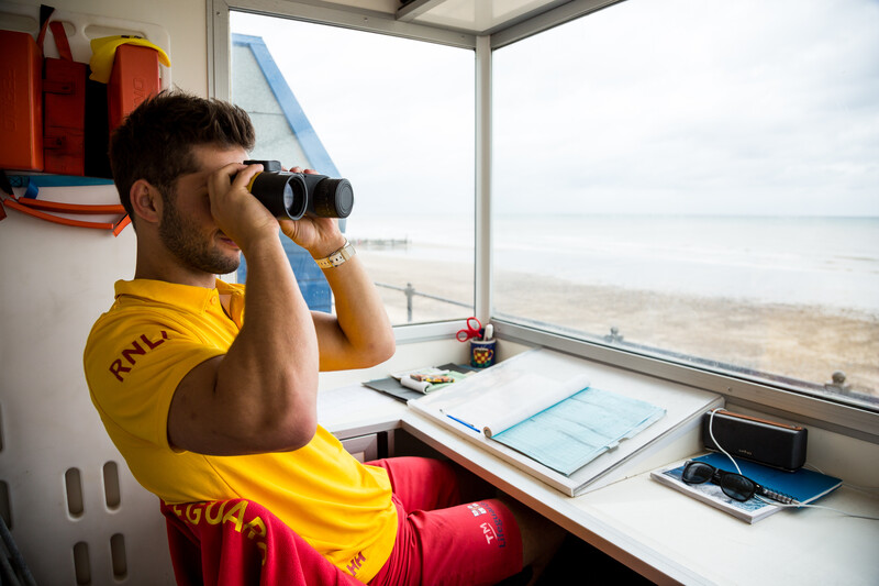 RNLI Lead Lifeguard Supervisor Ted Morgan on watch in yellow and red lifeguard uniform in lifeguard hut on beach