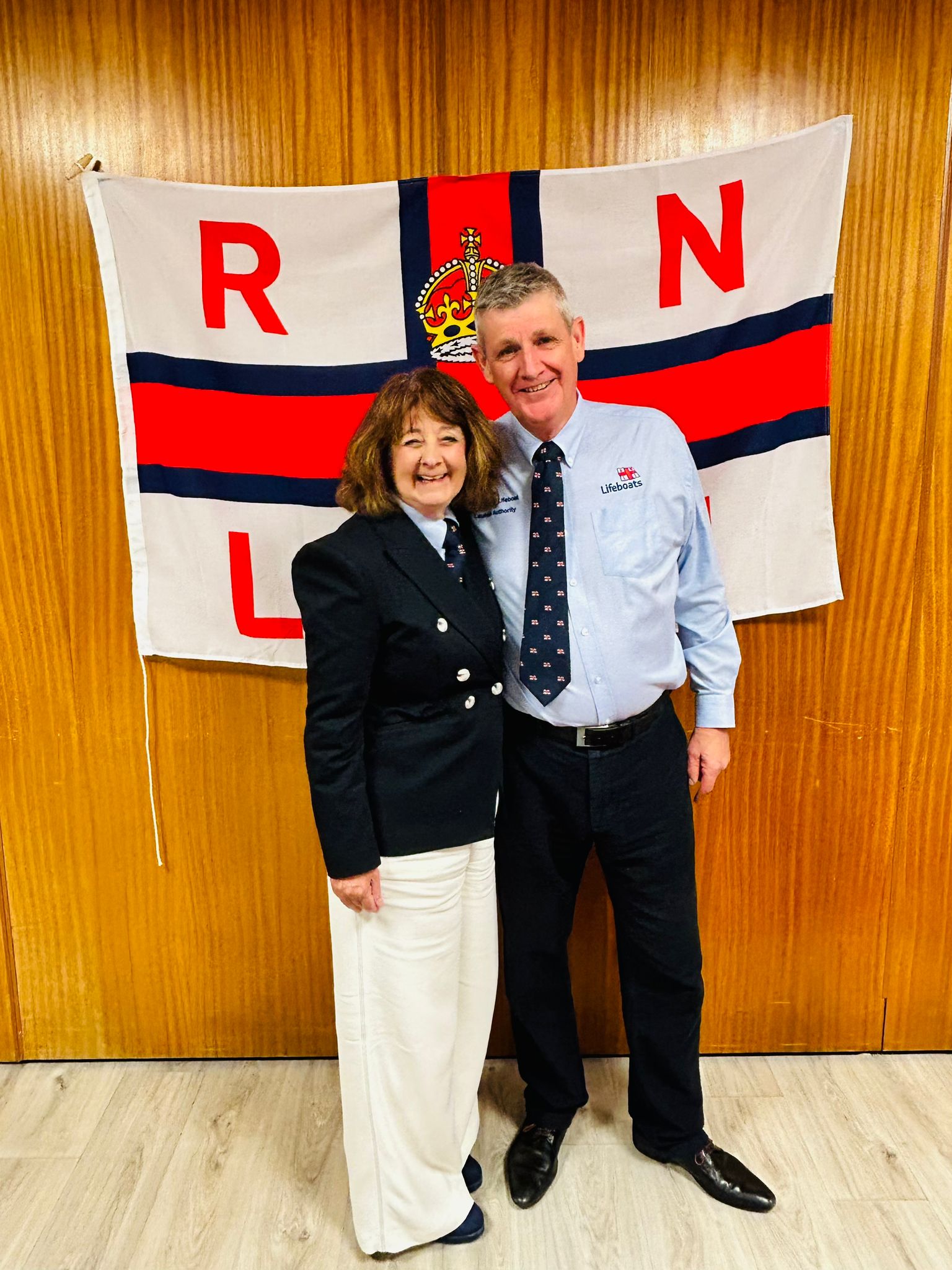 Barry Dock RNLI volunteers Avril and Bill Kitchen stood inf ront of RNLI flag.