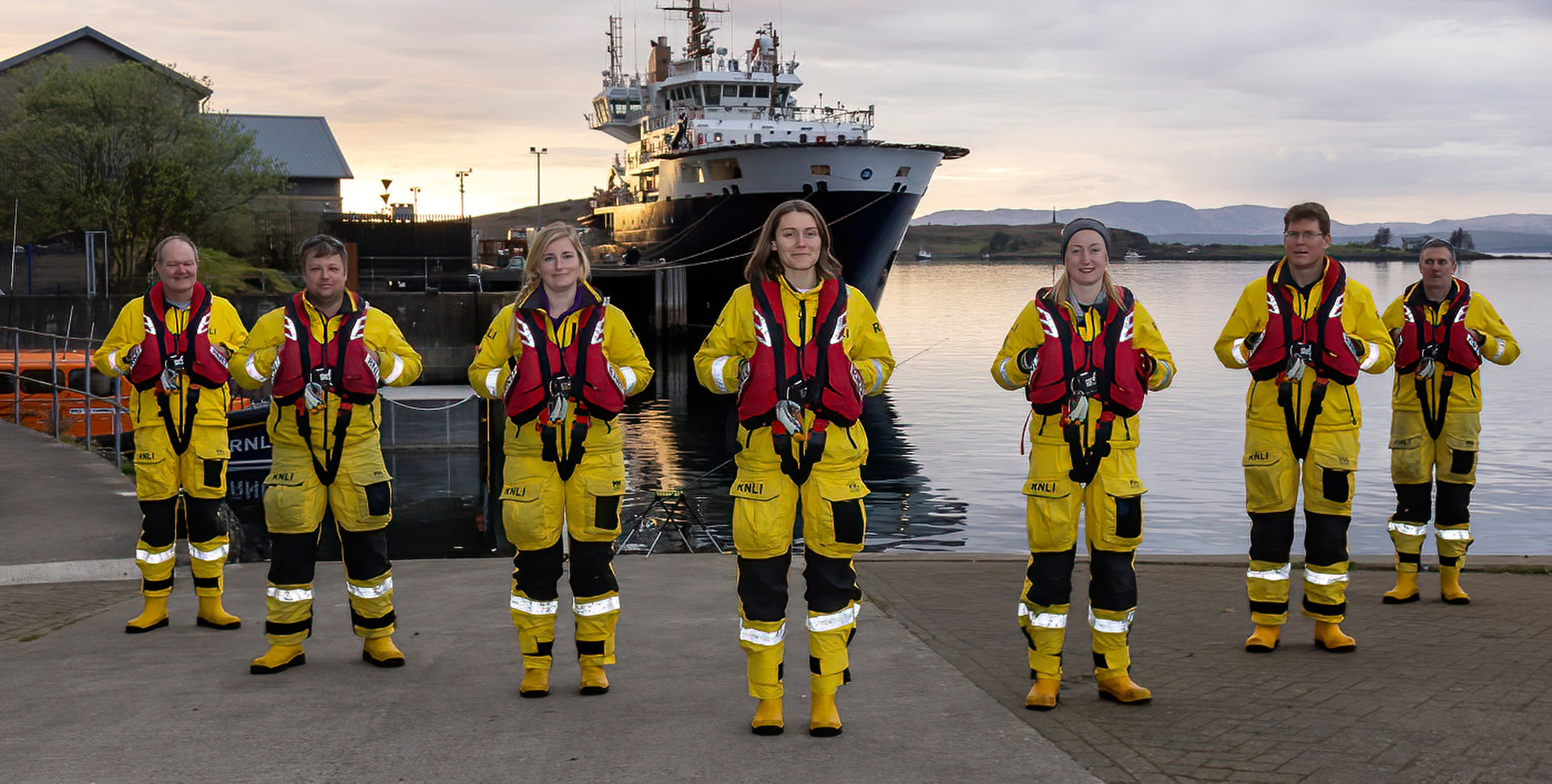 Seven members of the crew in full kit pose in the evening sunshine in front of the Oban lifeboat with the Northern Lighthouse vessel Pharos in the background