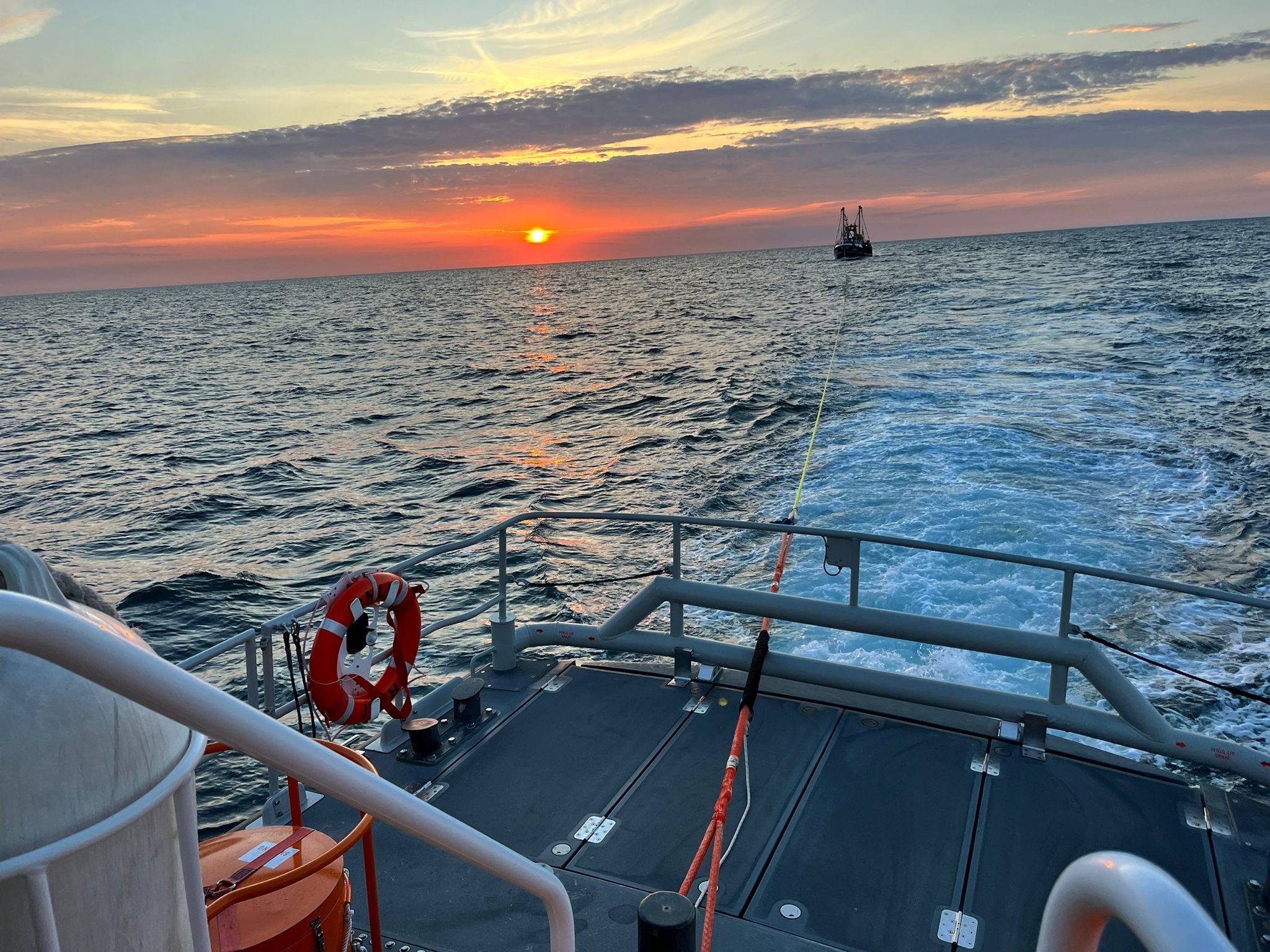 The image shows a beautiful sunset over the ocean with an orange and pink sky. In the foreground, there is a deck of a boat with safety equipment, including a life preserver and ropes. A tow line extends from this boat to another vessel visible in the distance.