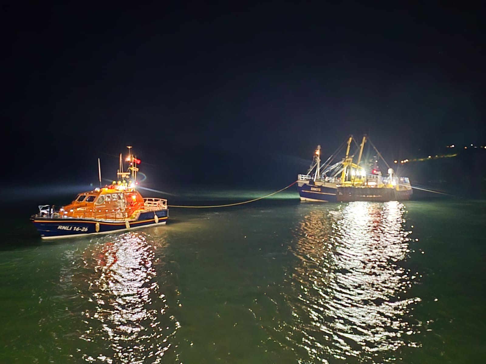 A nighttime scene showing a rescue boat and a fishing vessel on the water. The rescue boat, labeled 'RNLI 14-24,' is brightly lit and connected by a rope to the larger fishing vessel, which is also illuminated. The water reflects the lights from both boats.