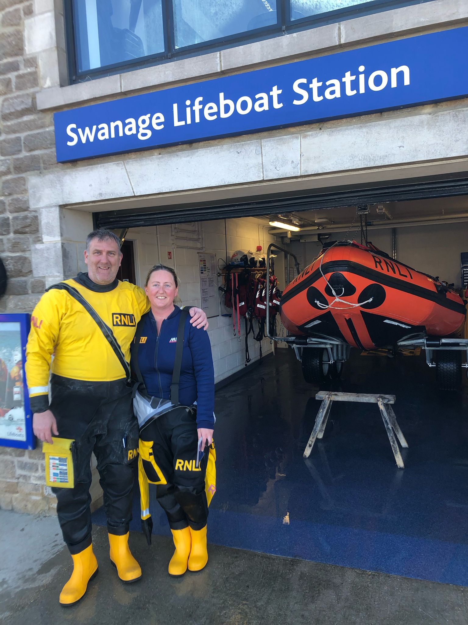 Man and woman wearing yellow and black drysuit gear stand in entryway to lifeboat station. Orange lifeboat is in background.