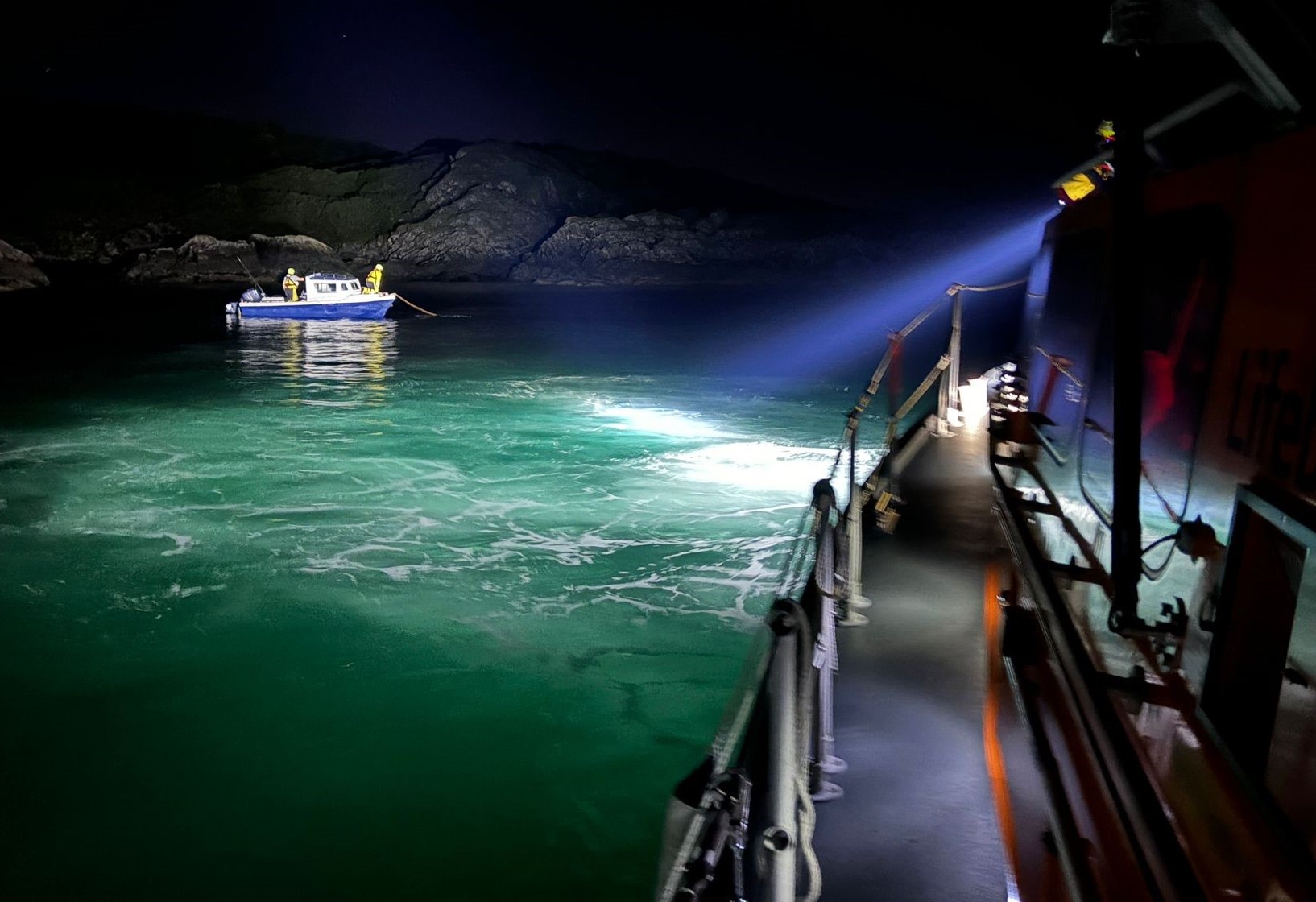 A searchlight from the Oban Lifeboat illuminates a small blue-hulled motorboat in the darkness with two lifeboat crew members visible onboard