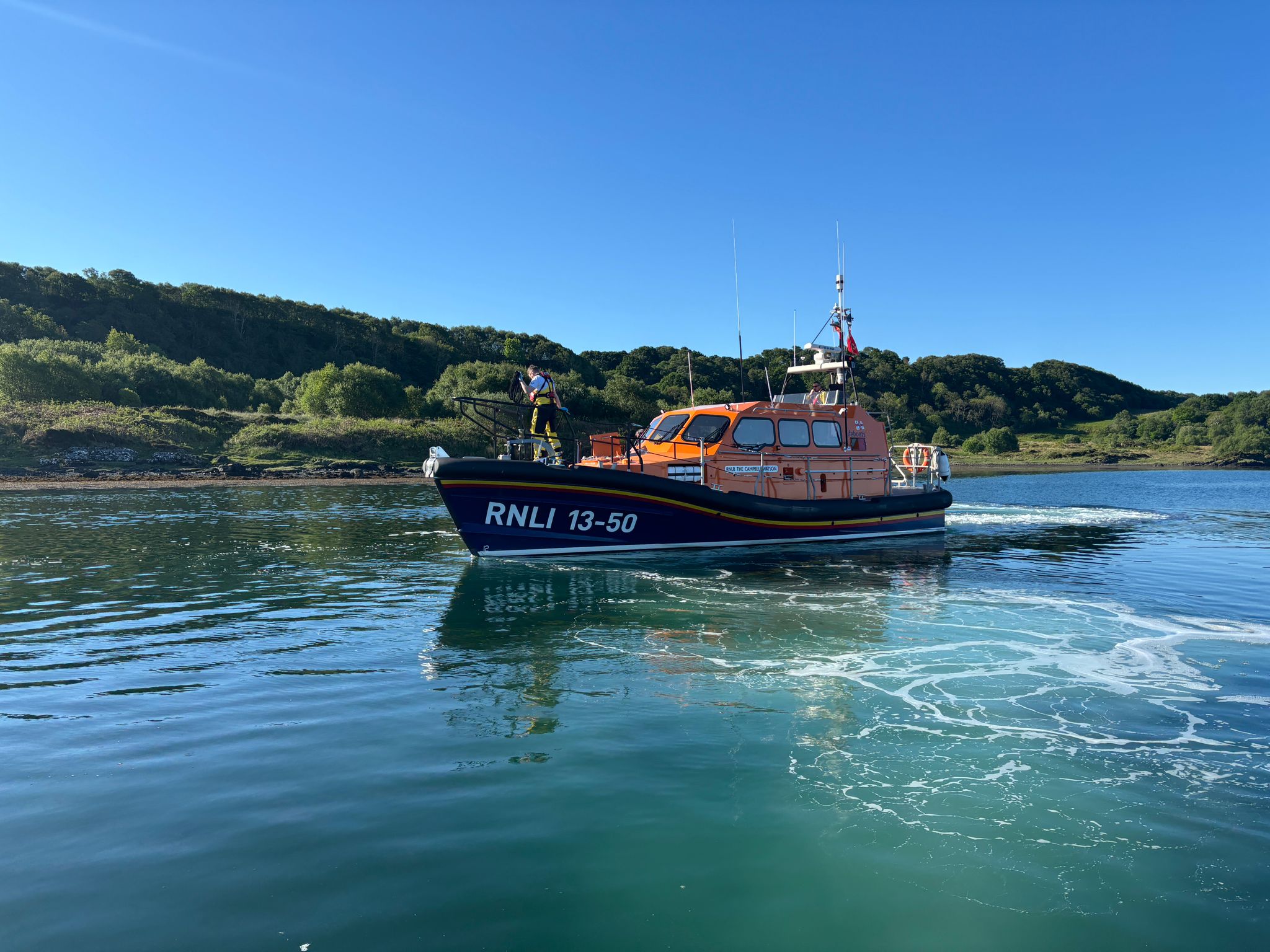 The Oban lifeboat in the morning sunshine, the calm waters frothed up as the lifeboat moves astern from the yacht.
