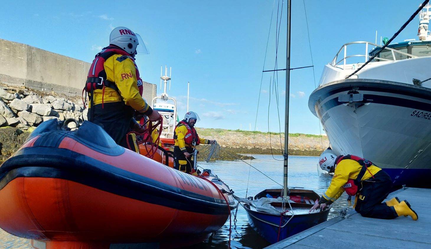 Two crew visible on the lifeboat on the water with a dinghy alongside coming in to dock. One crew member is on the pontoon securing the dinghy.