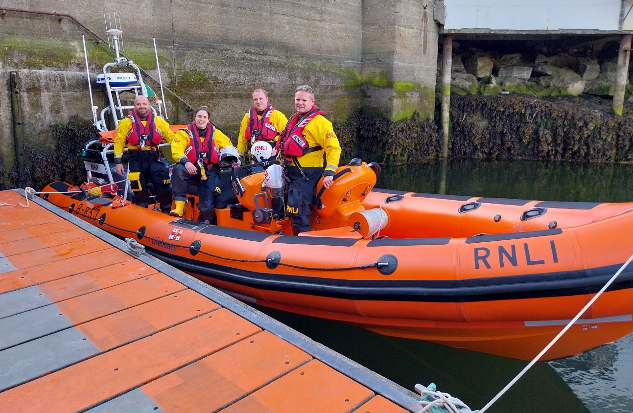 Four crew in drysuits on board the inshore lifeboat which is tied up at a pontoon. The four crew are holding their helmets and facing the camera