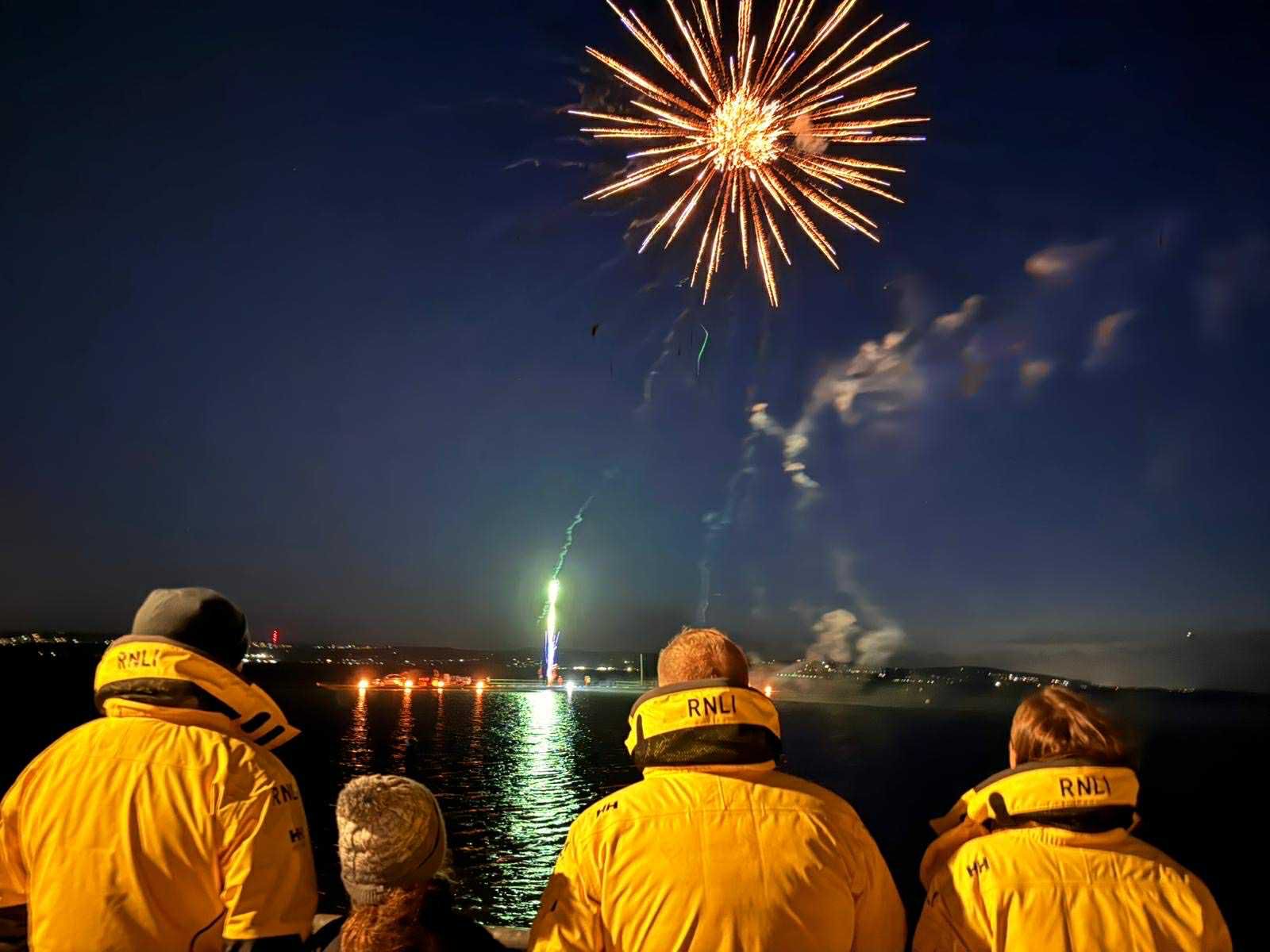 Three crew members in yellow coats can be seen looking out at the fireworks overhead.