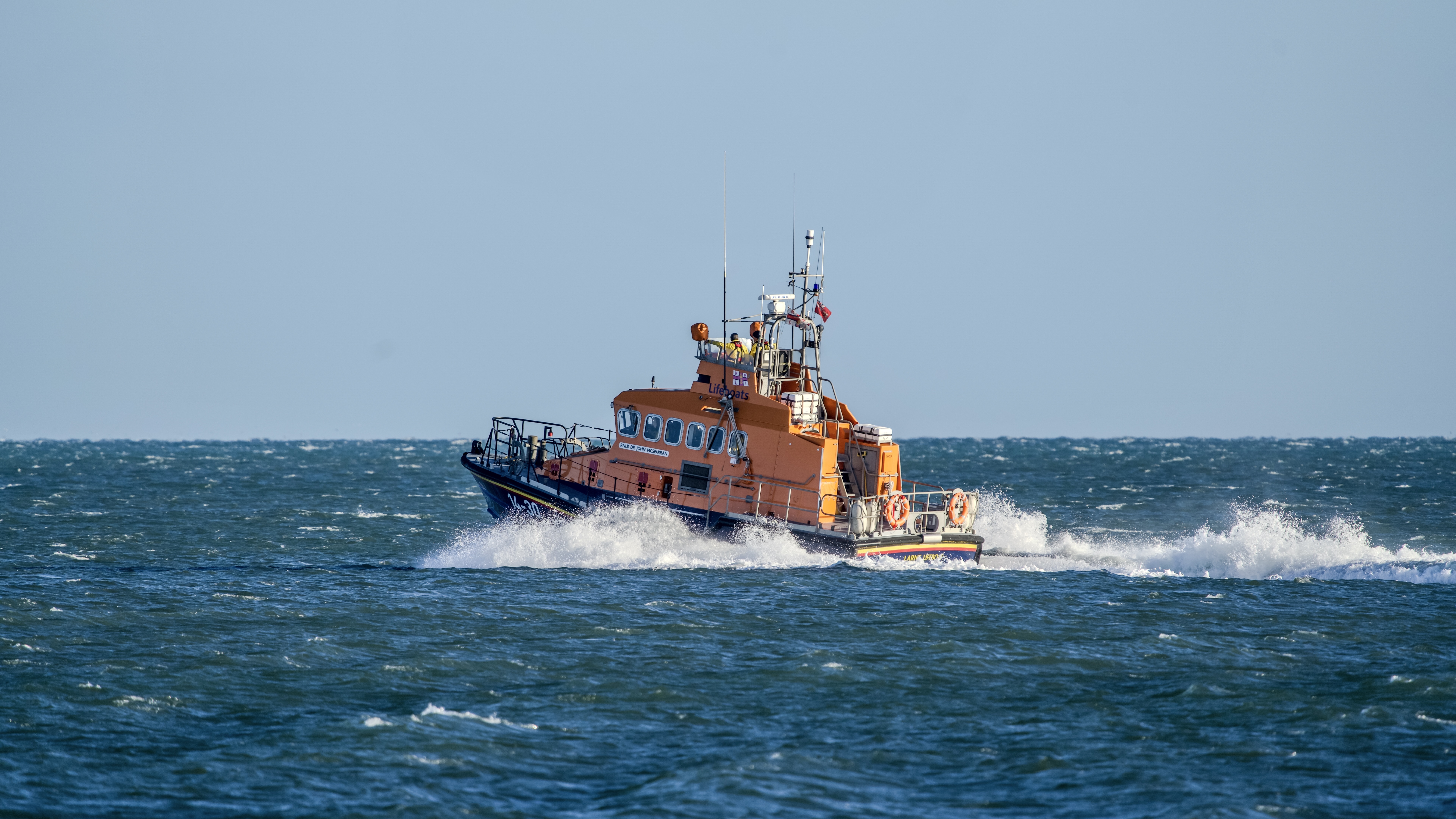 Larne Lifeboat speeding across the sea, creating waves and spray as it moves through the water. Weather conditions are clear with a blue sky and some moderate waves on the water