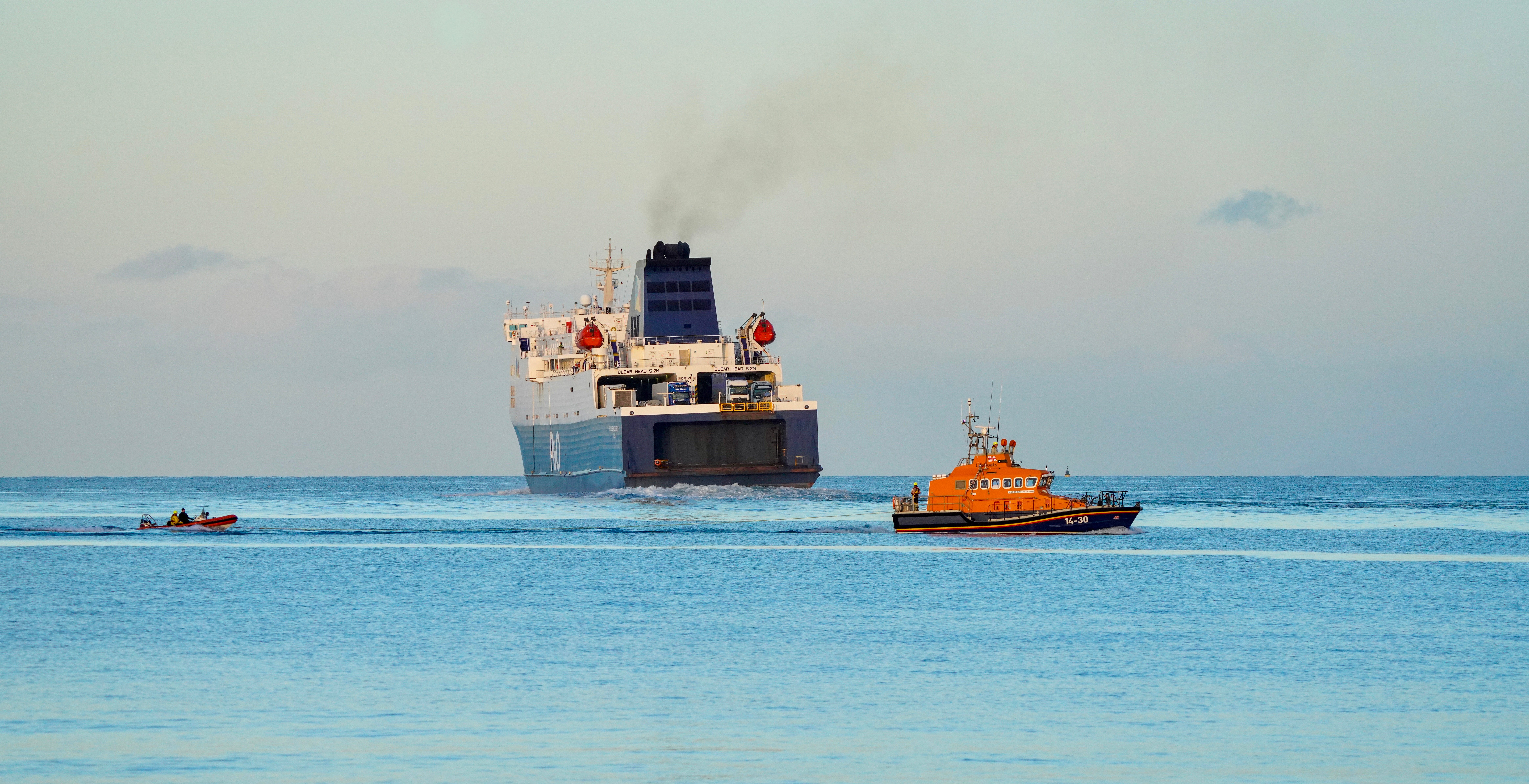 Larne’s all-weather lifeboat tows a small inflatable boat across calm waters as a passenger ferry heads out toward the horizon.