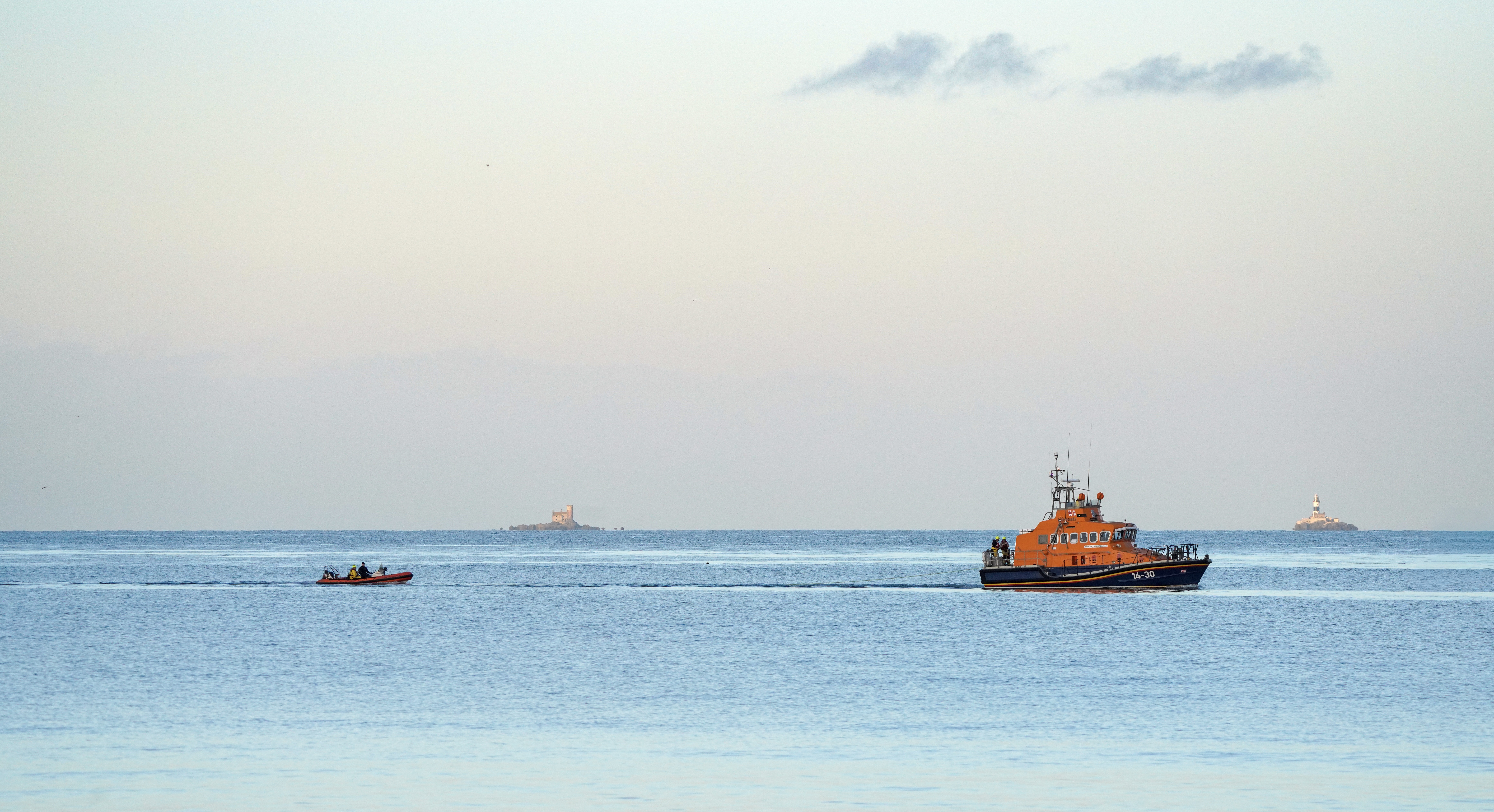 Larne’s all-weather lifeboat tows a small inflatable boat across calm waters; the Maidens Lighthouses are in the background