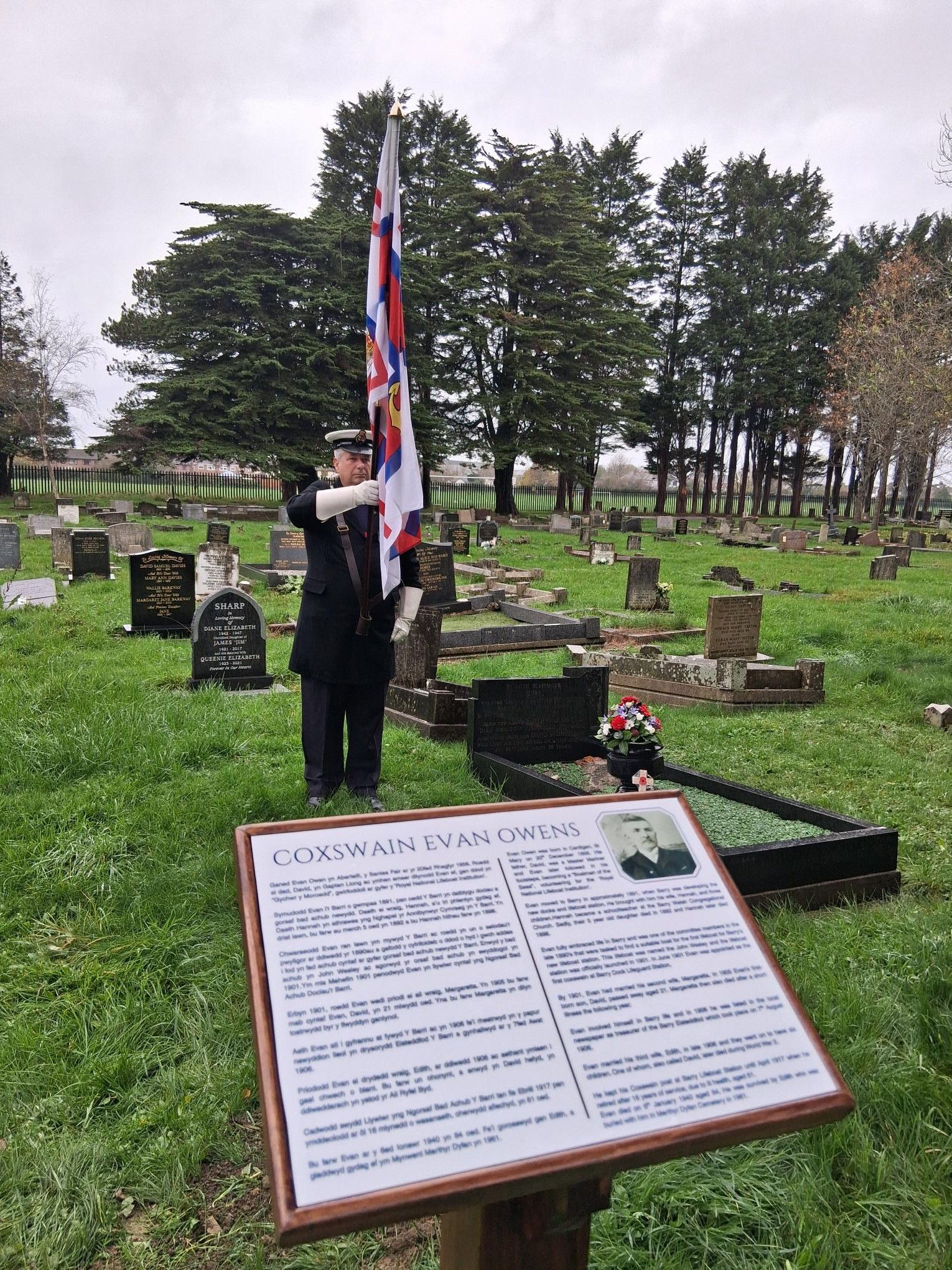 The image has the plaque in the foreground and an RNLI volunteer in uniform holding the RNLI flag
