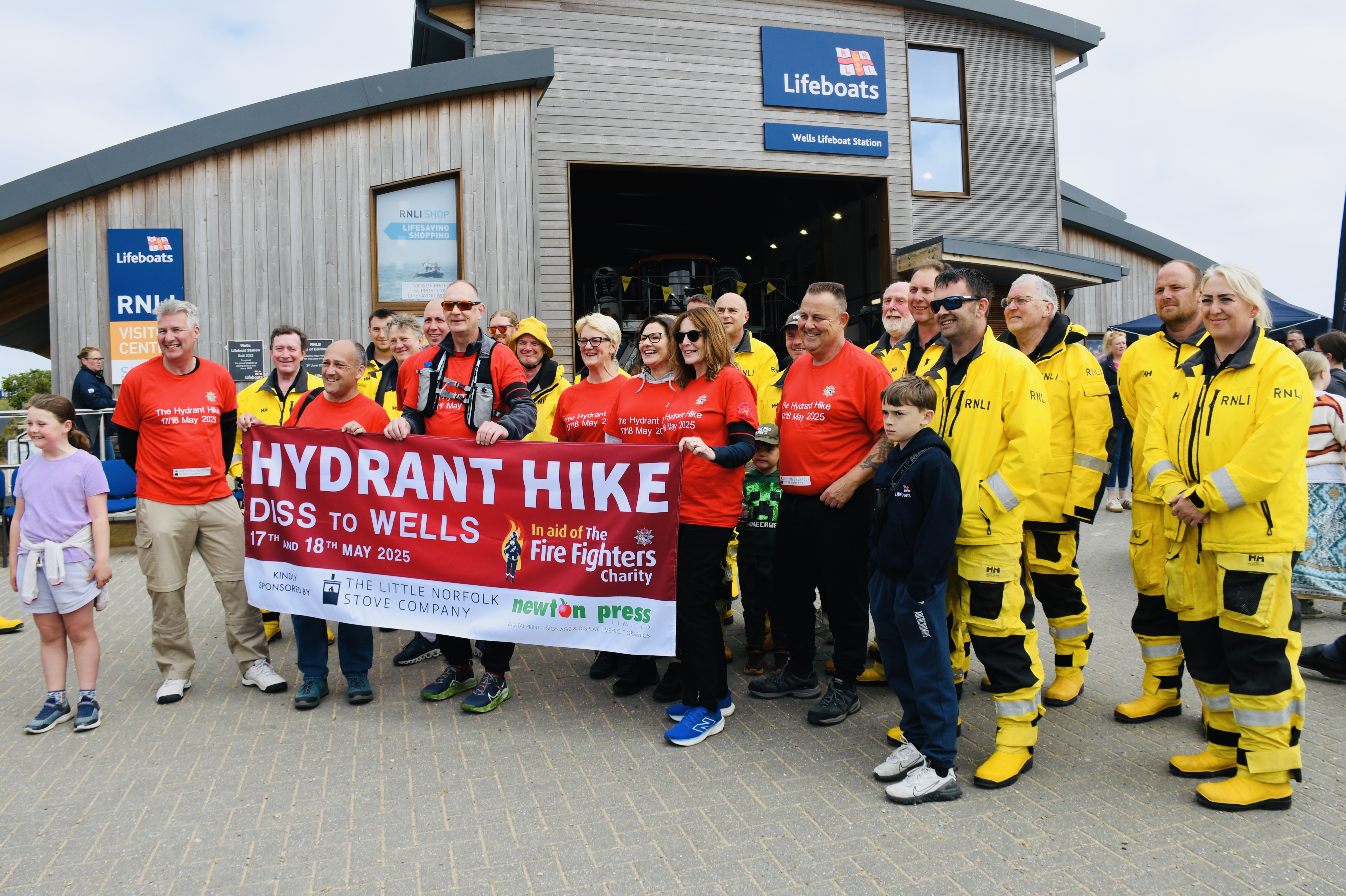 Wells RNLI crew members in full yellow kit with Norfolk fire and rescue members in red holding a hydrant hike sign at Mayday event station in the background