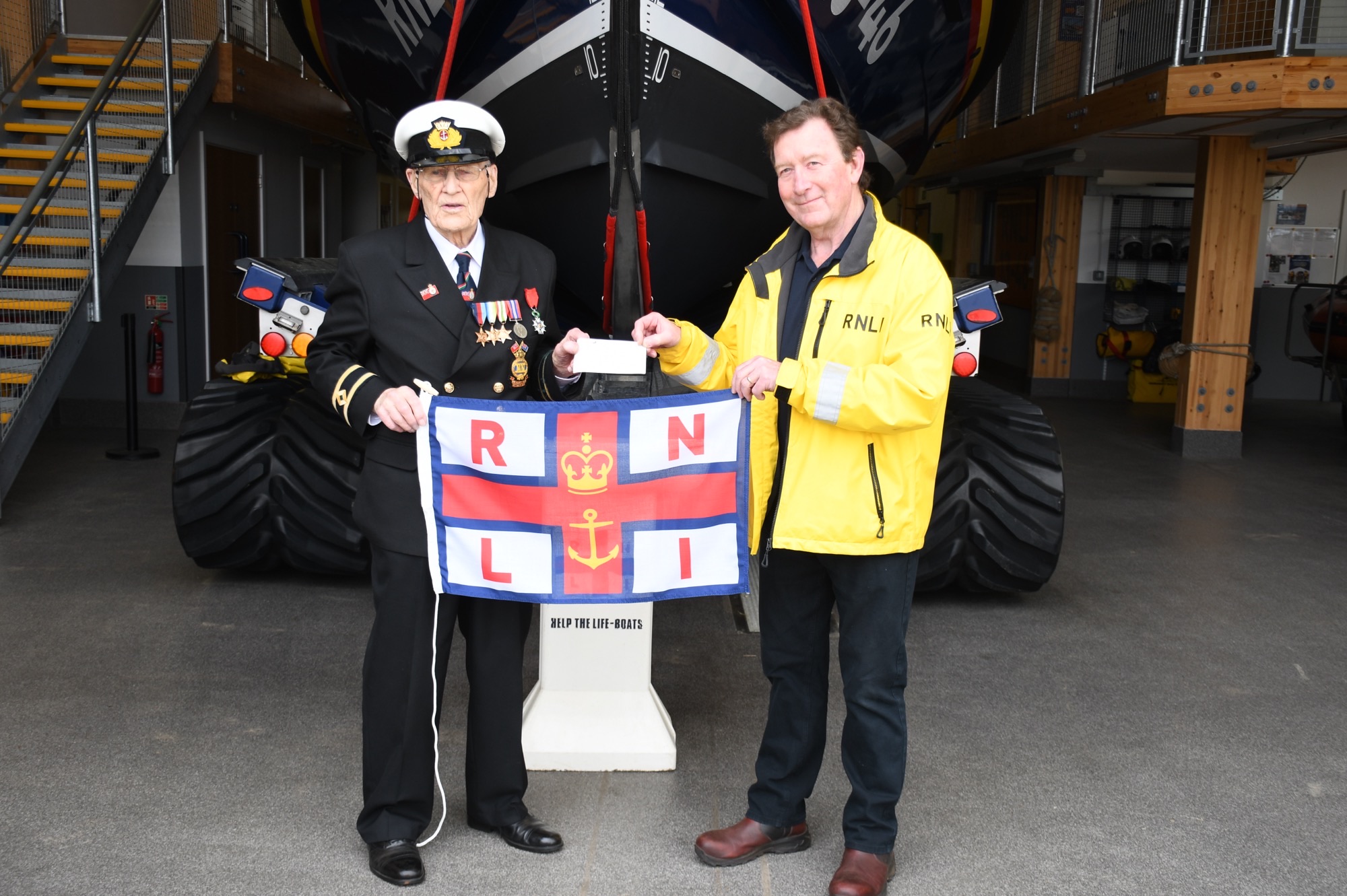 Ronald Quested in full navy uniform presenting donation to Wells RNLI lifeboats operations manager Chris Hardy in front of lifeboat holding an RNLI flag