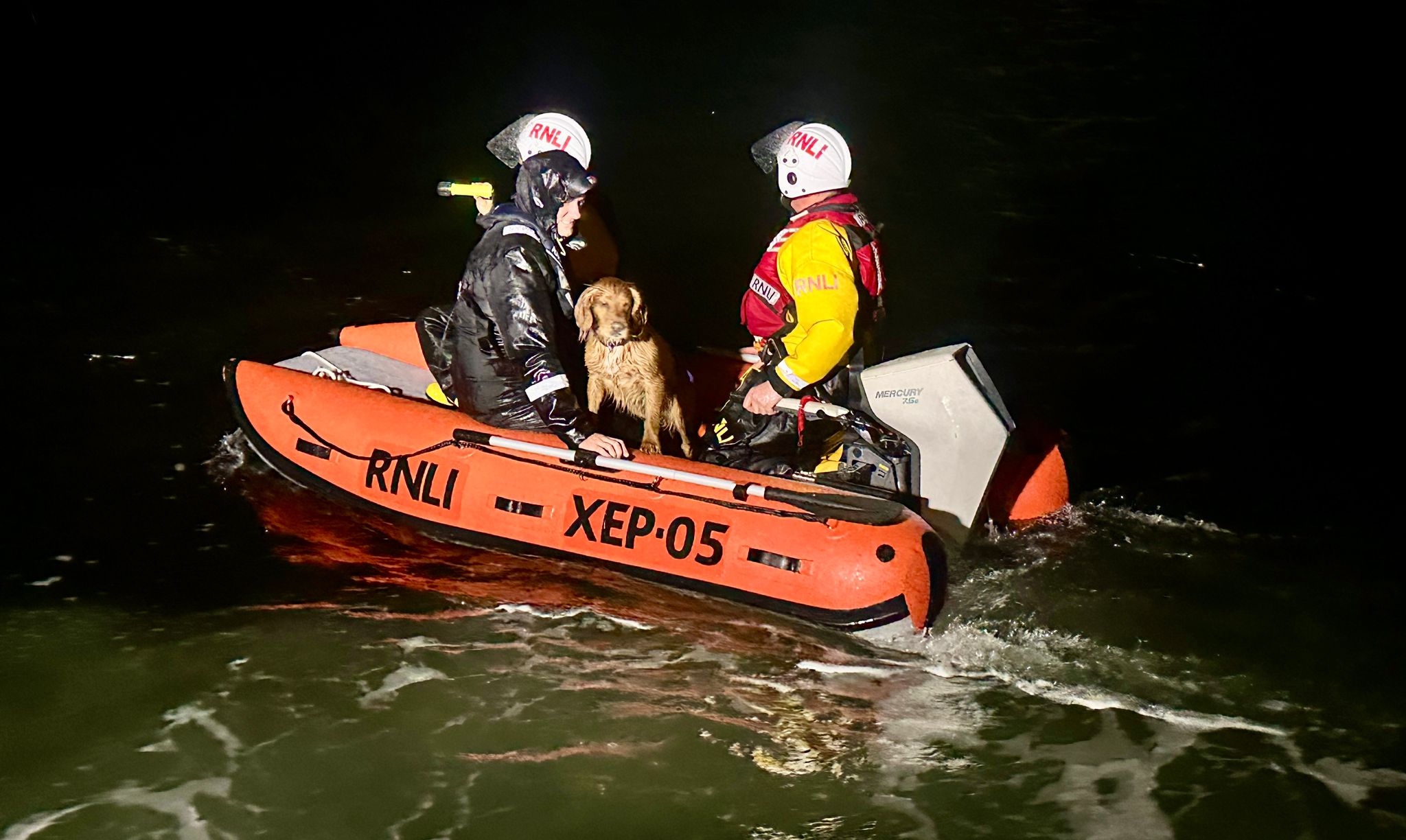Two lifeboat crew in yellows fit into a small inflatable craft alongside a man in a black coat and the golden cocker spaniel.
