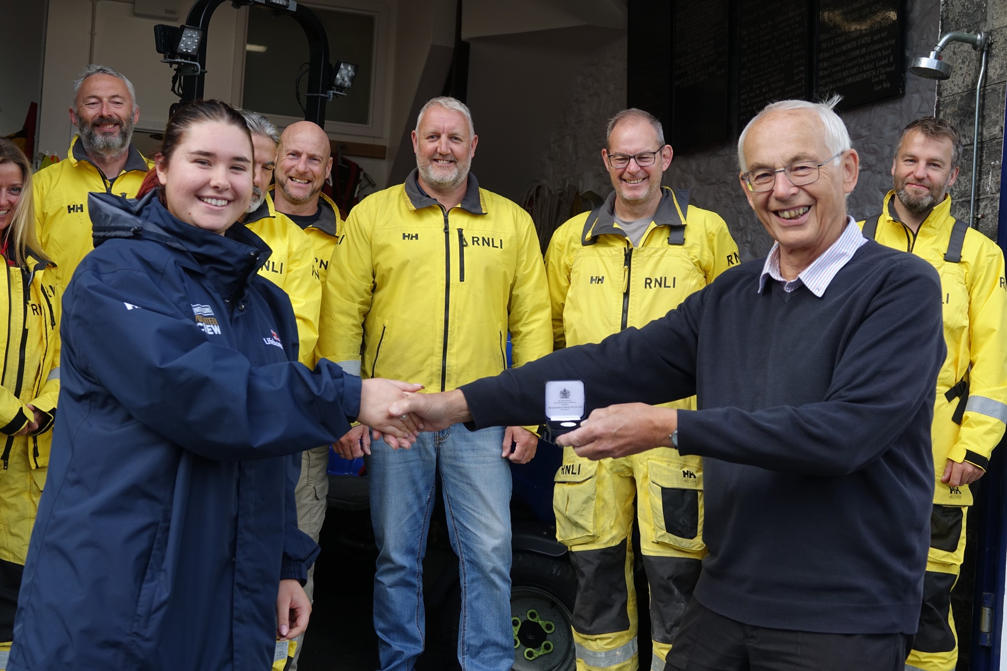 Ruth and Ken shaking hands and ken holding his medal with crew in background.
