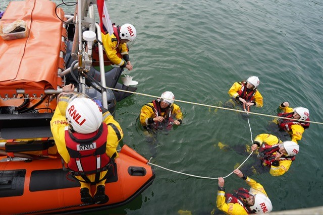 People in RNLI drysuits and helmets gather at the stern of a lifeboat, in the water