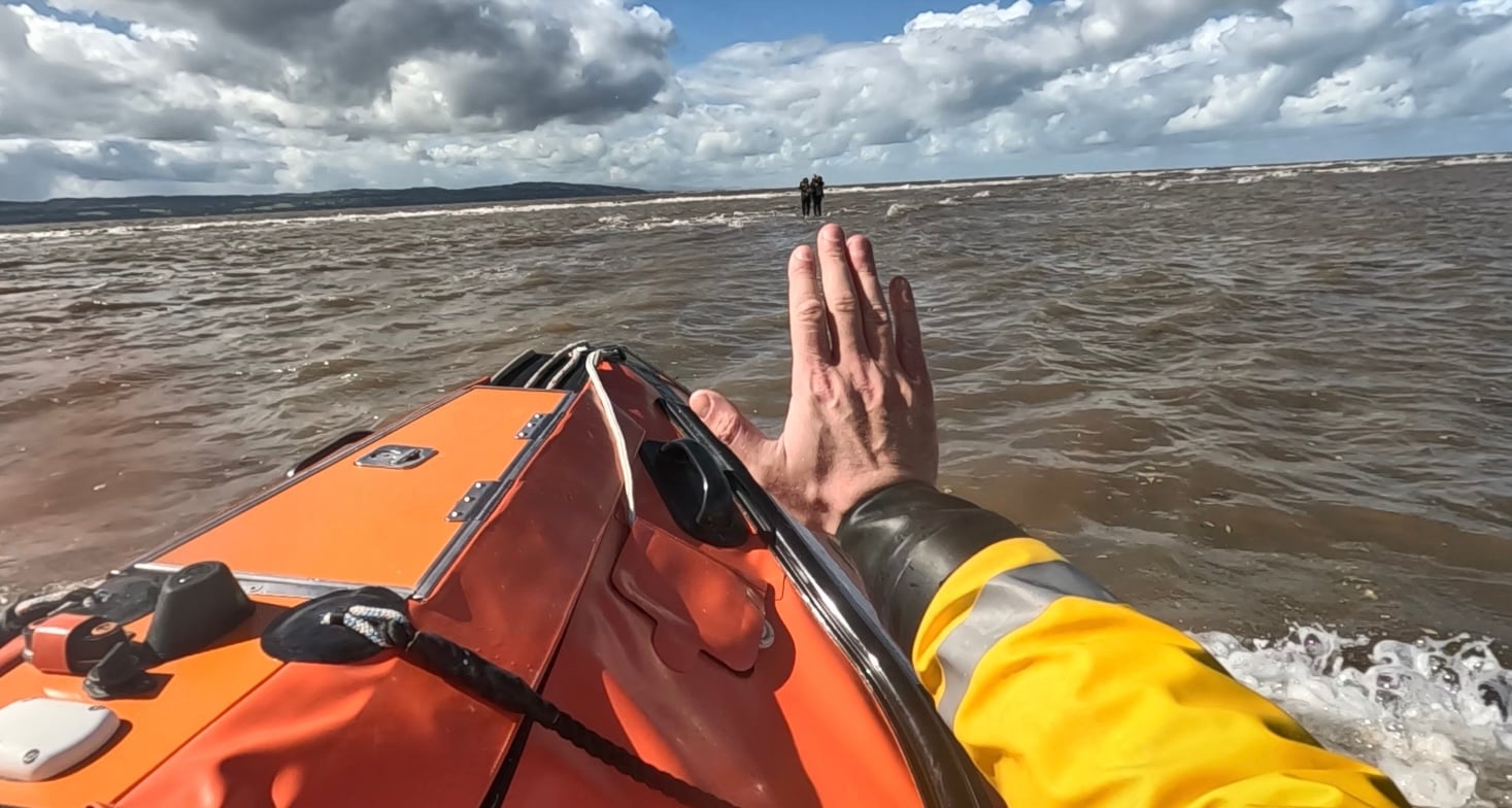 The lifeboat can be seen approaching two individuals stood on rocks and completely surrounded by water while a crew member signals with a wave to them.