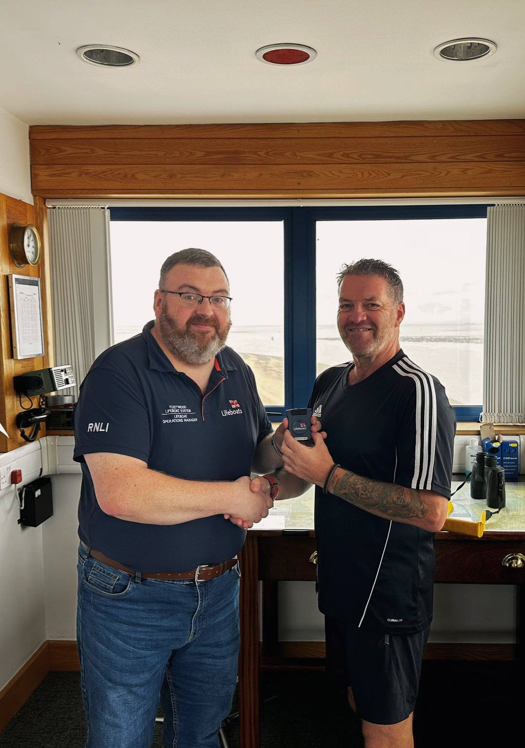Two men indoors, handing over an RNLI crew pager. A window behind shows a view of Morecambe Bay.