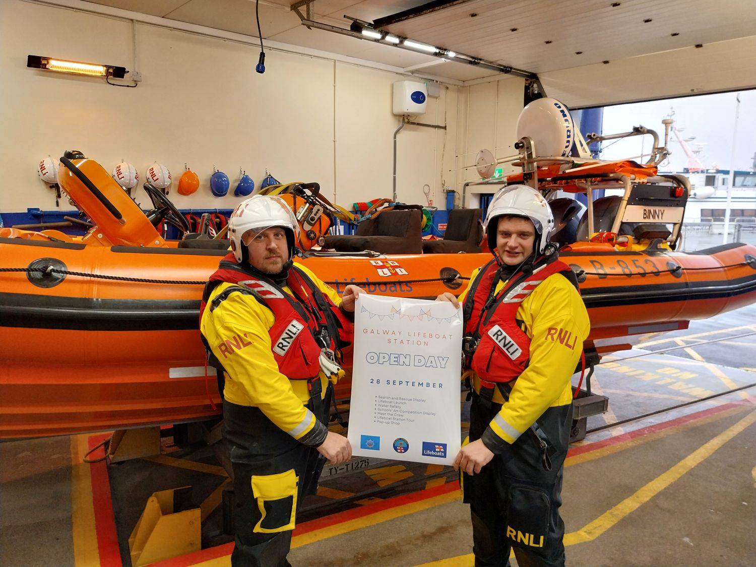 Two crew in drysuits and lifejackets in the boat house holding an open day poster while standing in front of the inshore lifeboat.