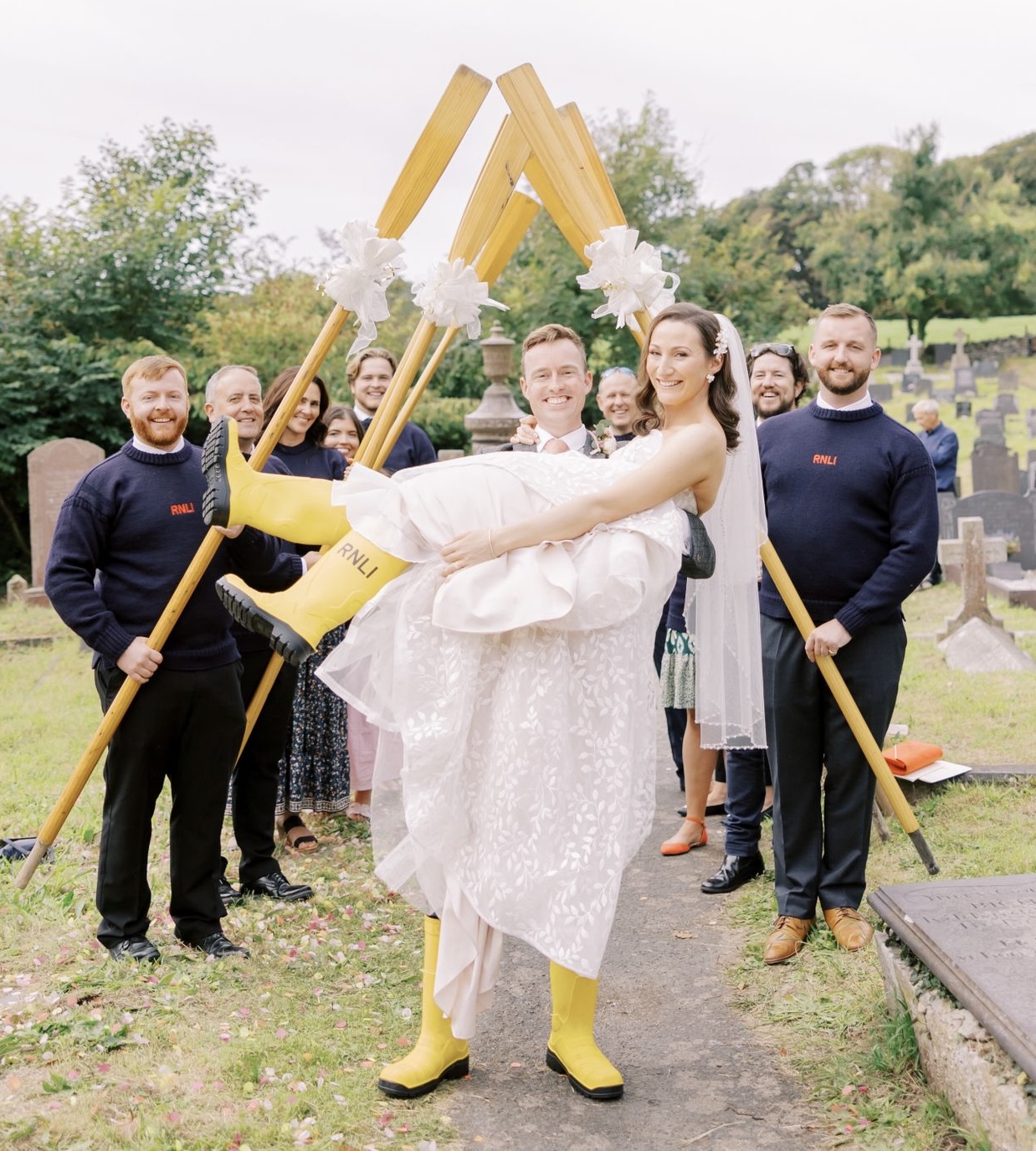 The groom lifts the bride up while they both wear the RNLI yellow wellies in front of the crew with the guard of honour.