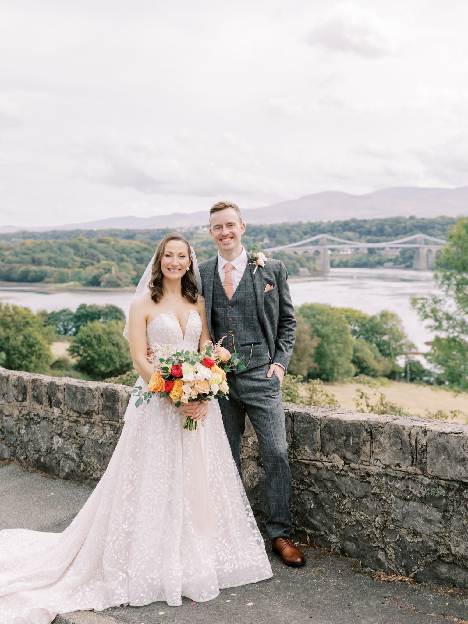 The couple stand in front of the Menai Bridge in the background 