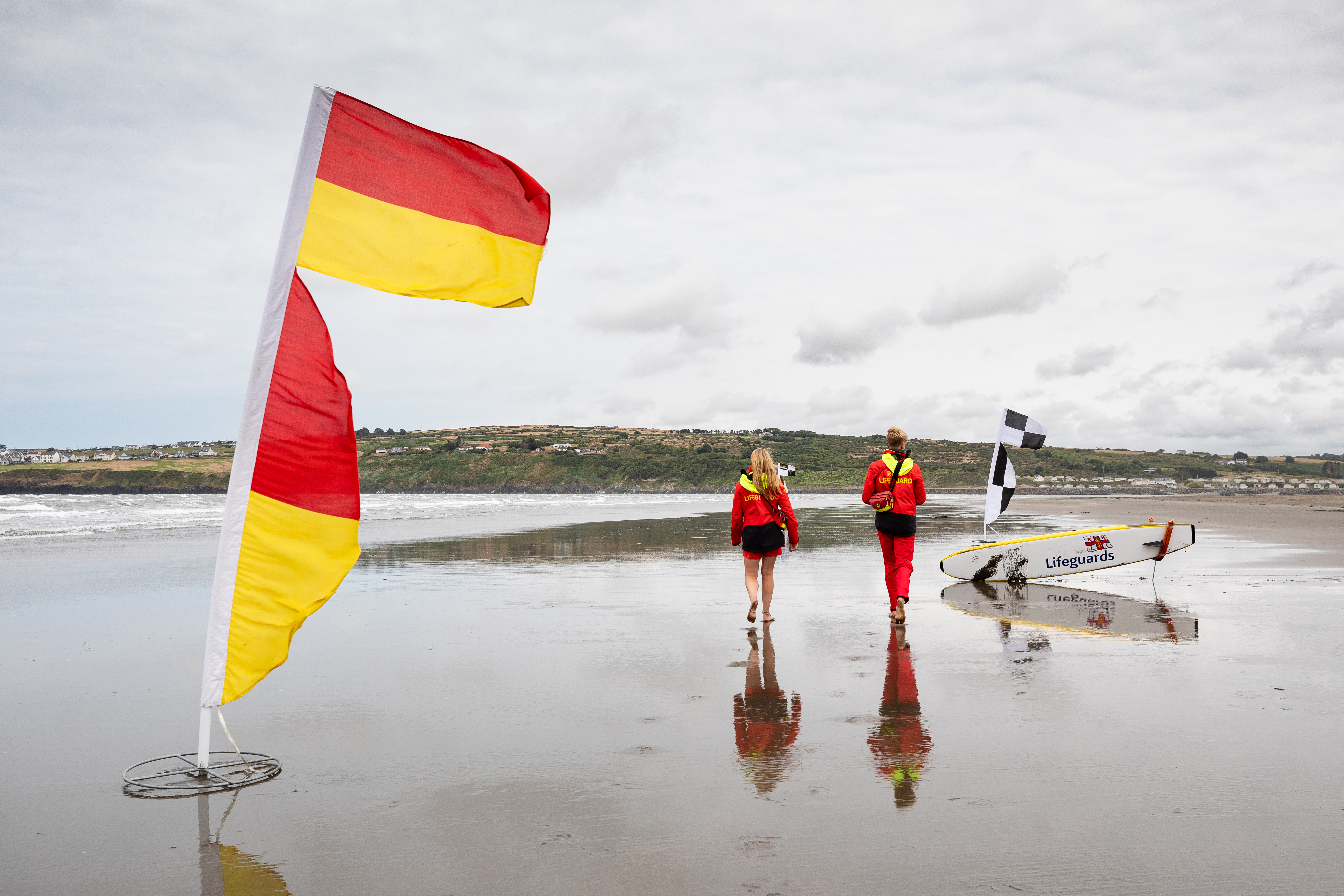Two RNLI lifeguards walk on a beach towards a surfboard away from red and yellow flags
