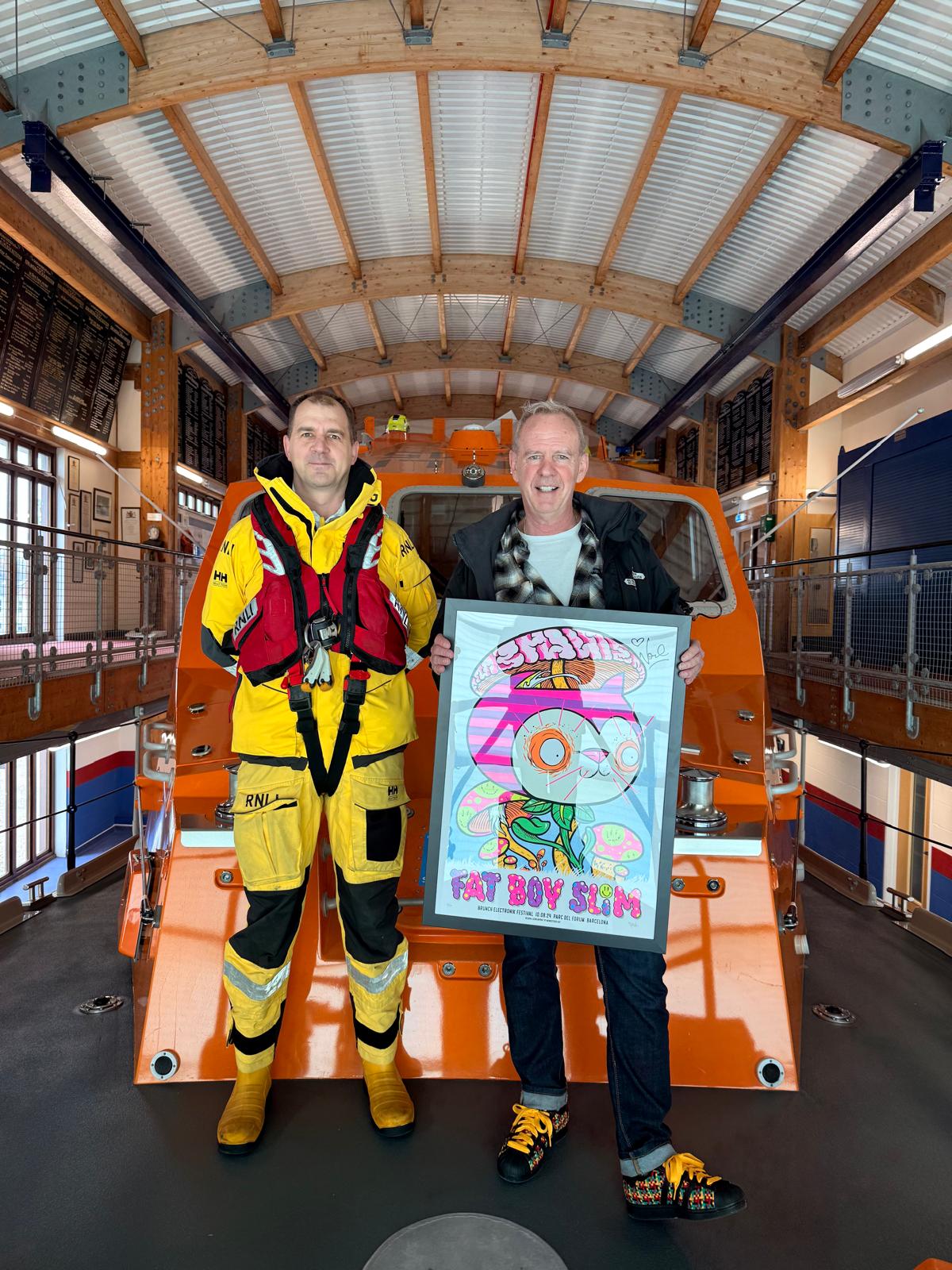 DJ Fatboy Slim is pictured on the front of the ALB with Shoreham RNLI coxswain, Simon Williams. He is holding a poster and smiling.