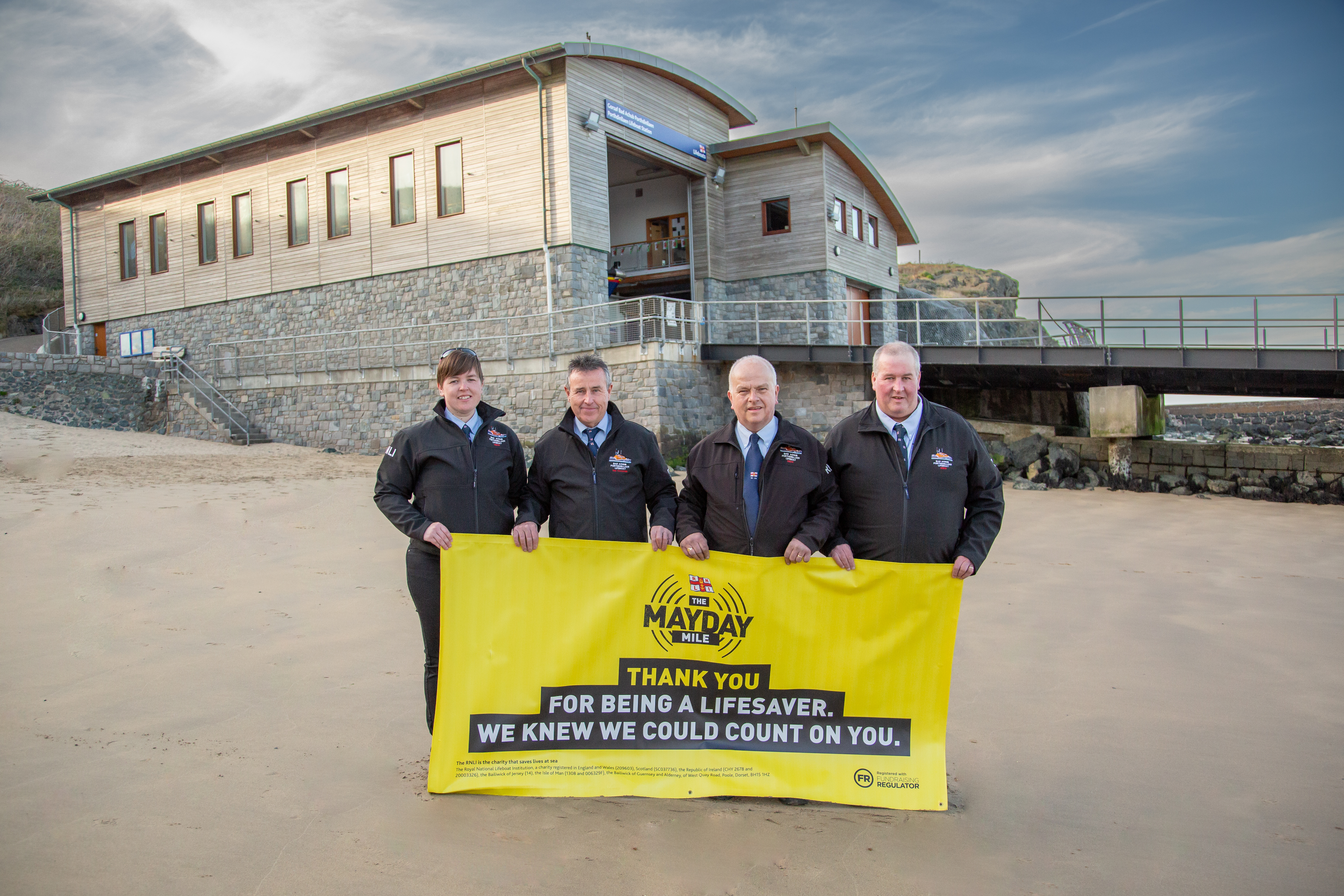 Landscape image of the four volunteers holding Mayday banner with a backdrop of Porthdinllaen lifeboat station.